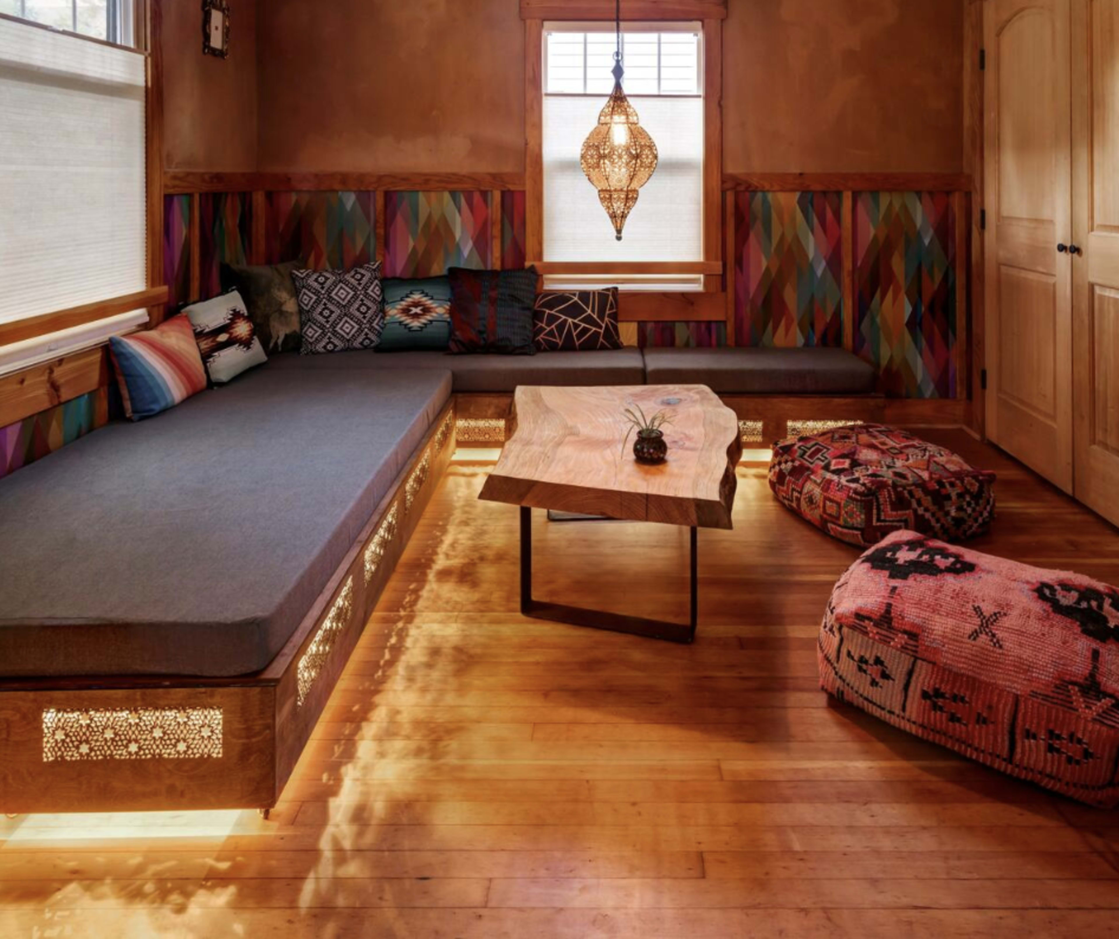 A cozy sitting area with a low wooden coffee table, a cushioned bench against a colorful wall, and patterned poufs on a wooden floor.