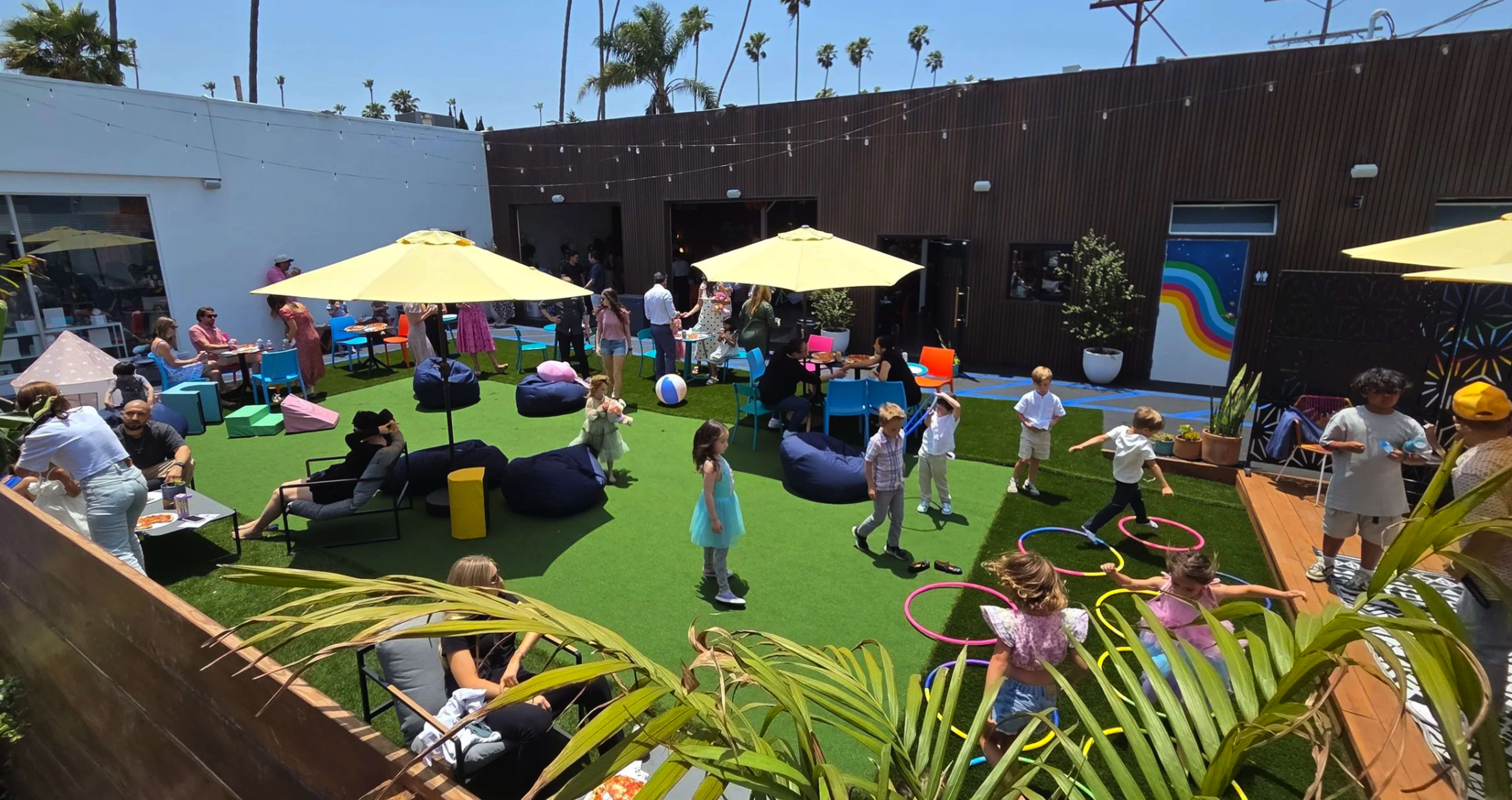 A vibrant outdoor space features children playing on green turf surrounded by colorful umbrellas, bean bags, and various activities, with adults mingling in the background.