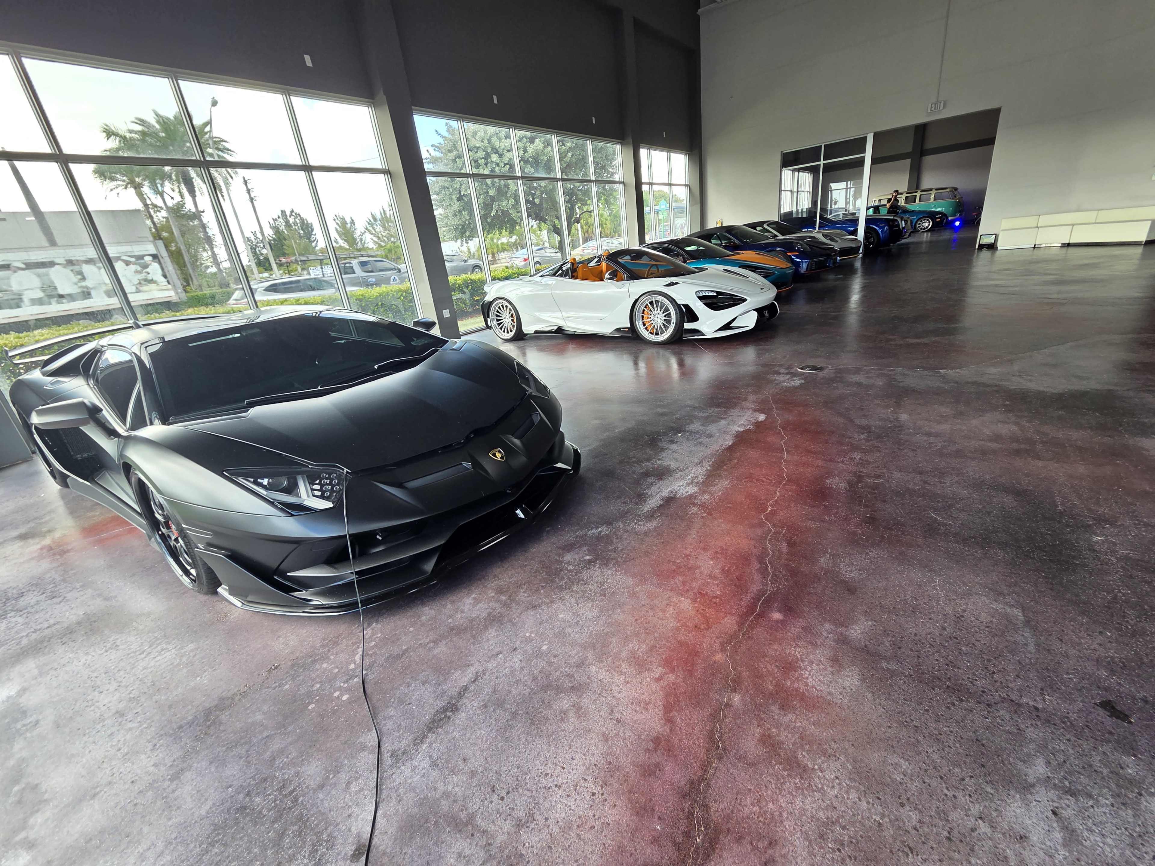 A row of luxury cars, including a black Lamborghini and a white McLaren, are displayed in a well-lit showroom with large windows.