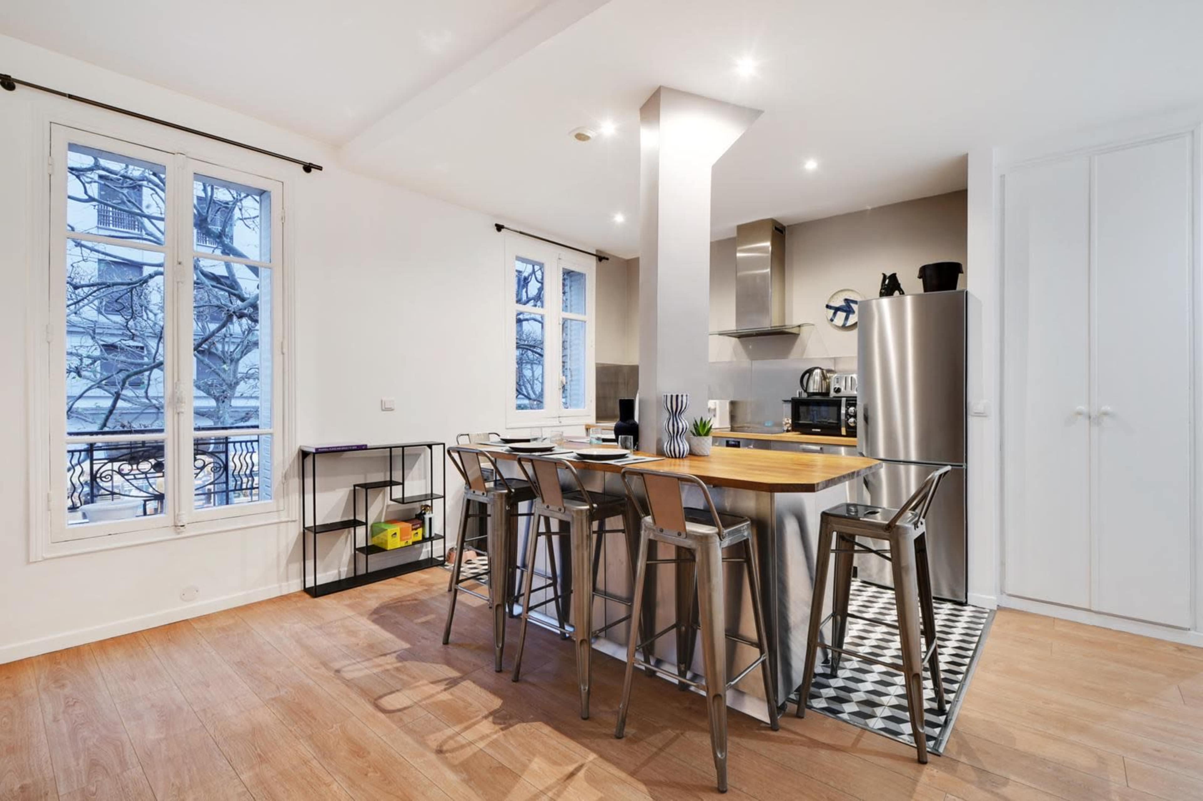 A modern kitchen with a central island, high stools, stainless steel appliances, and large windows overlooking a balcony.