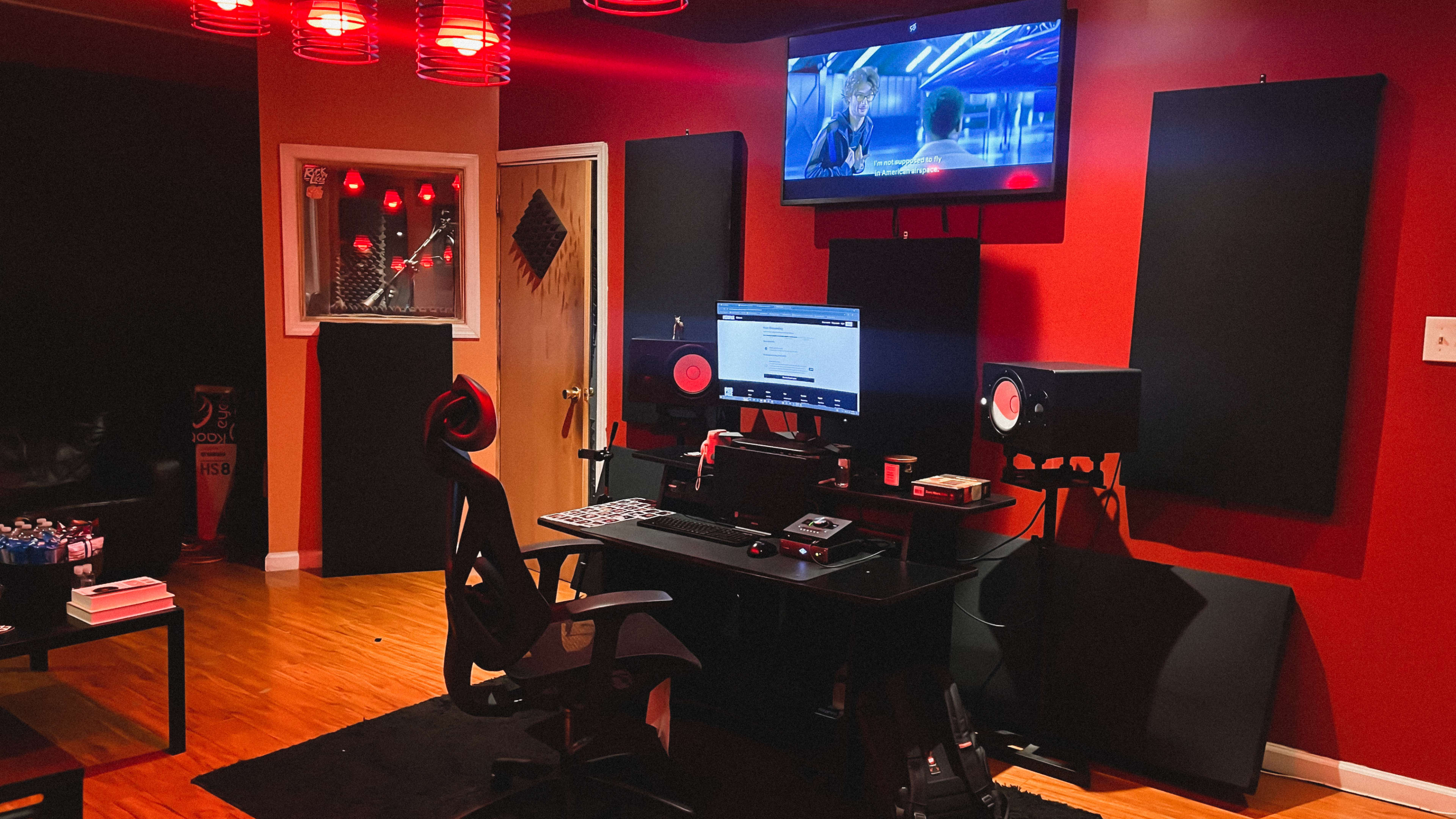 The image shows a recording studio setup with a computer desk, audio equipment, and red accent lighting in a dark room.