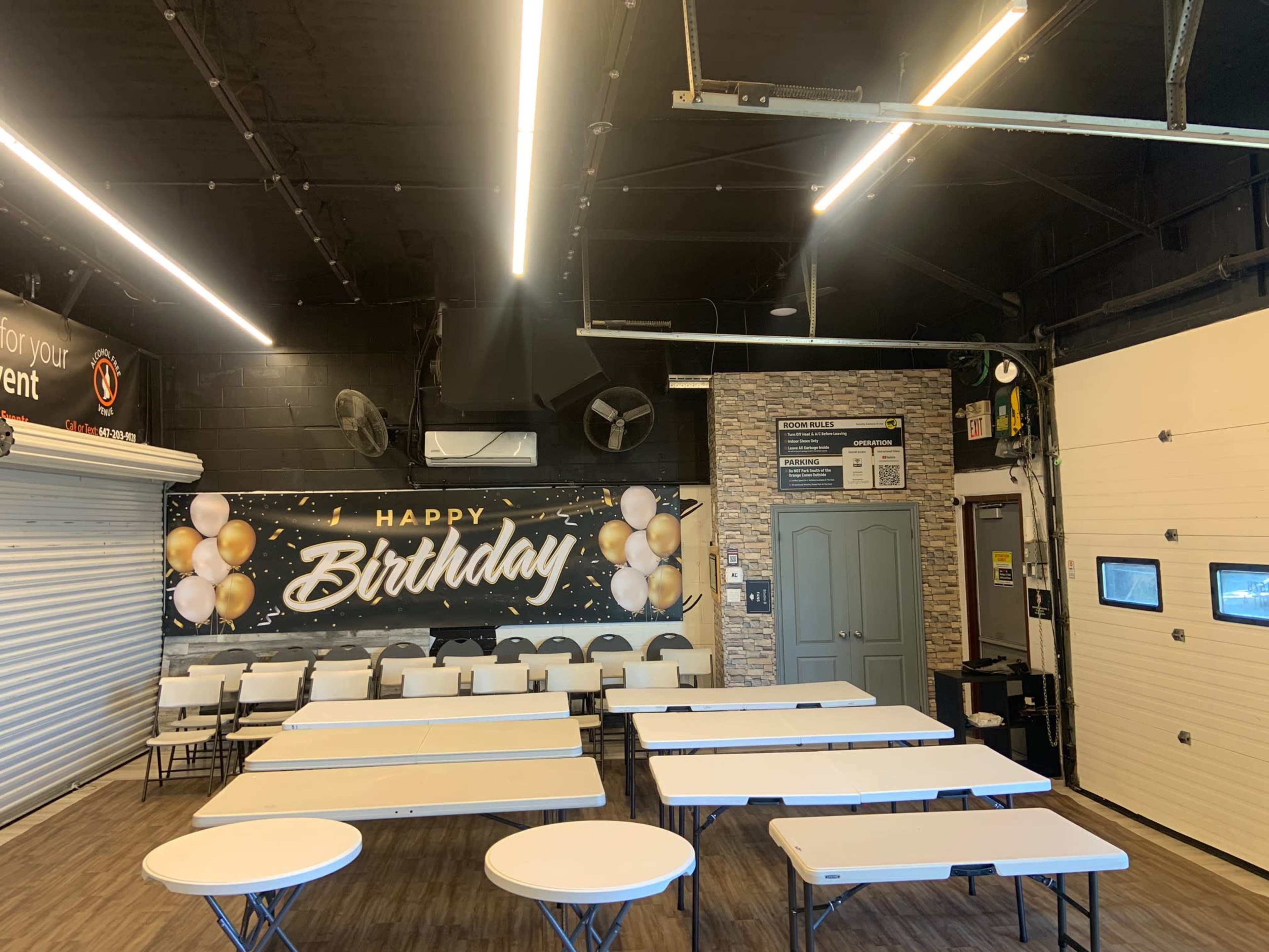 The image shows an indoor event space decorated with a "Happy Birthday" banner, featuring tables arranged in a row and a garage door in the background.