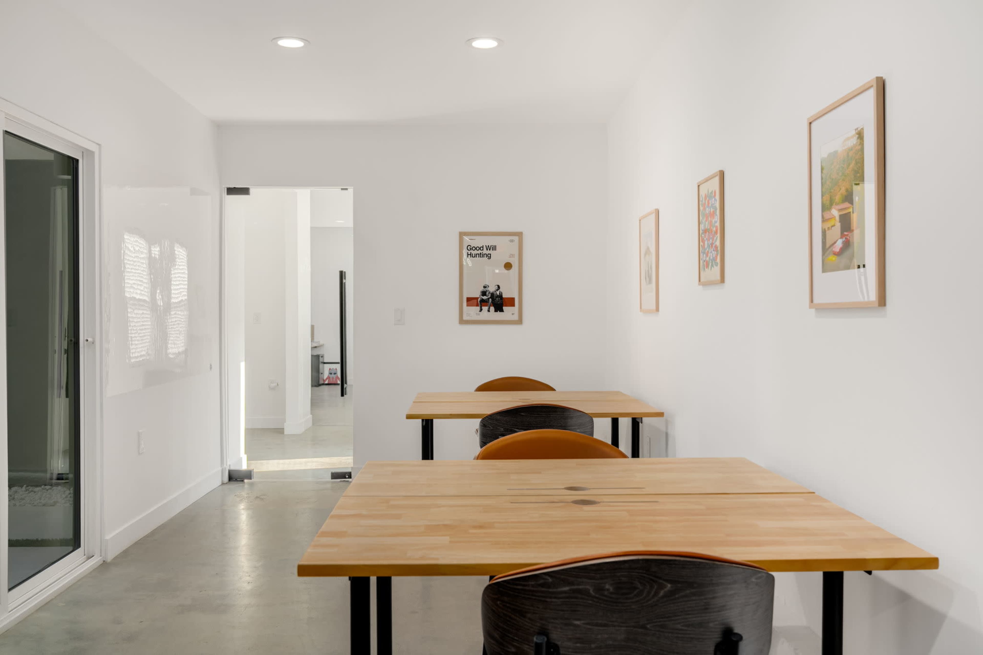 The image shows a bright, minimalist room with three wooden tables and framed artworks on the white walls.
