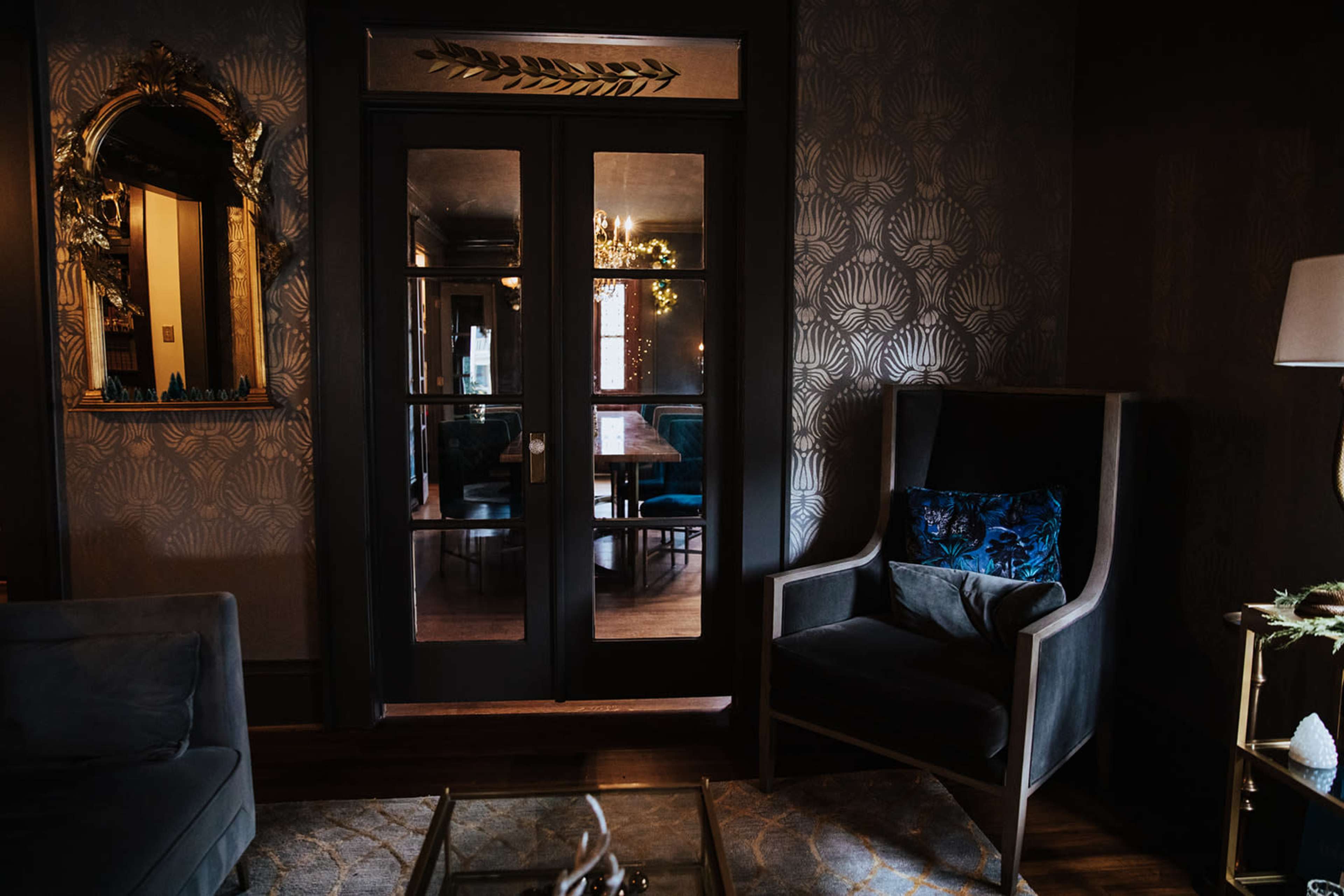 A stylish room features a plush armchair beside a glass coffee table and a set of double doors leading to a space with a chandelier reflecting on the walls.
