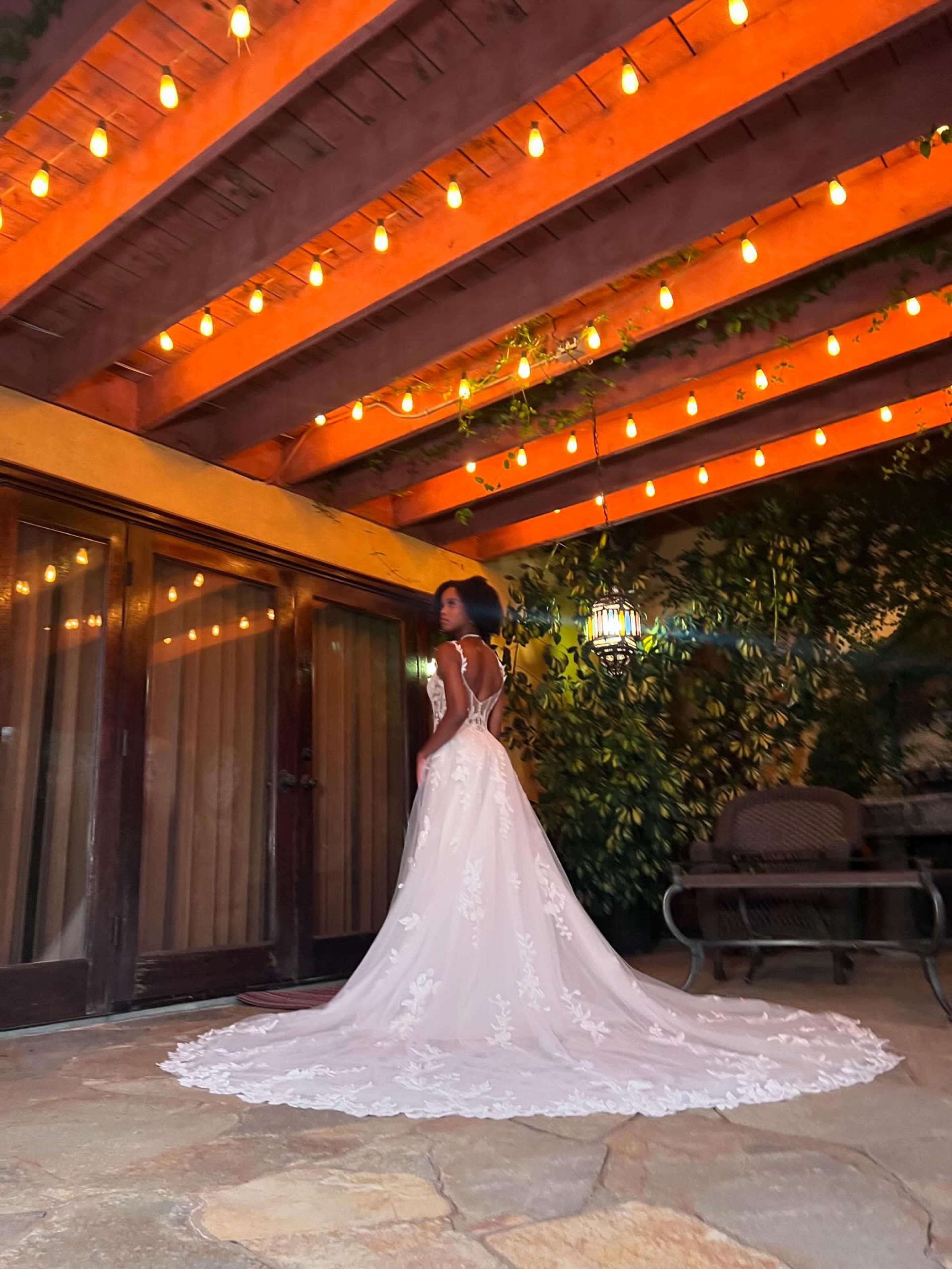 A woman in a white wedding dress stands gracefully in a courtyard illuminated by string lights.