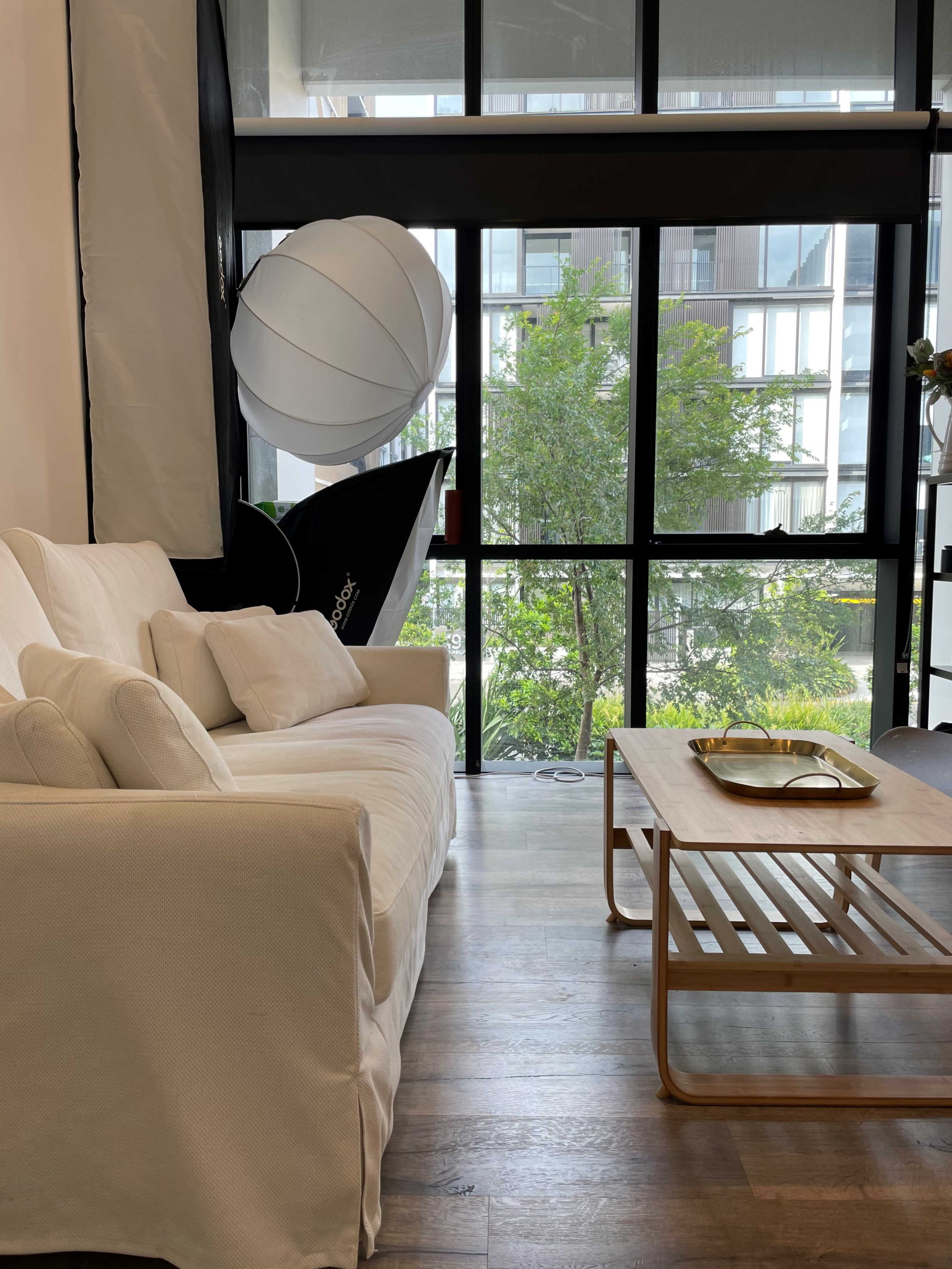 A white couch with decorative pillows sits beside a wooden coffee table, facing large windows that overlook greenery and an outdoor space.