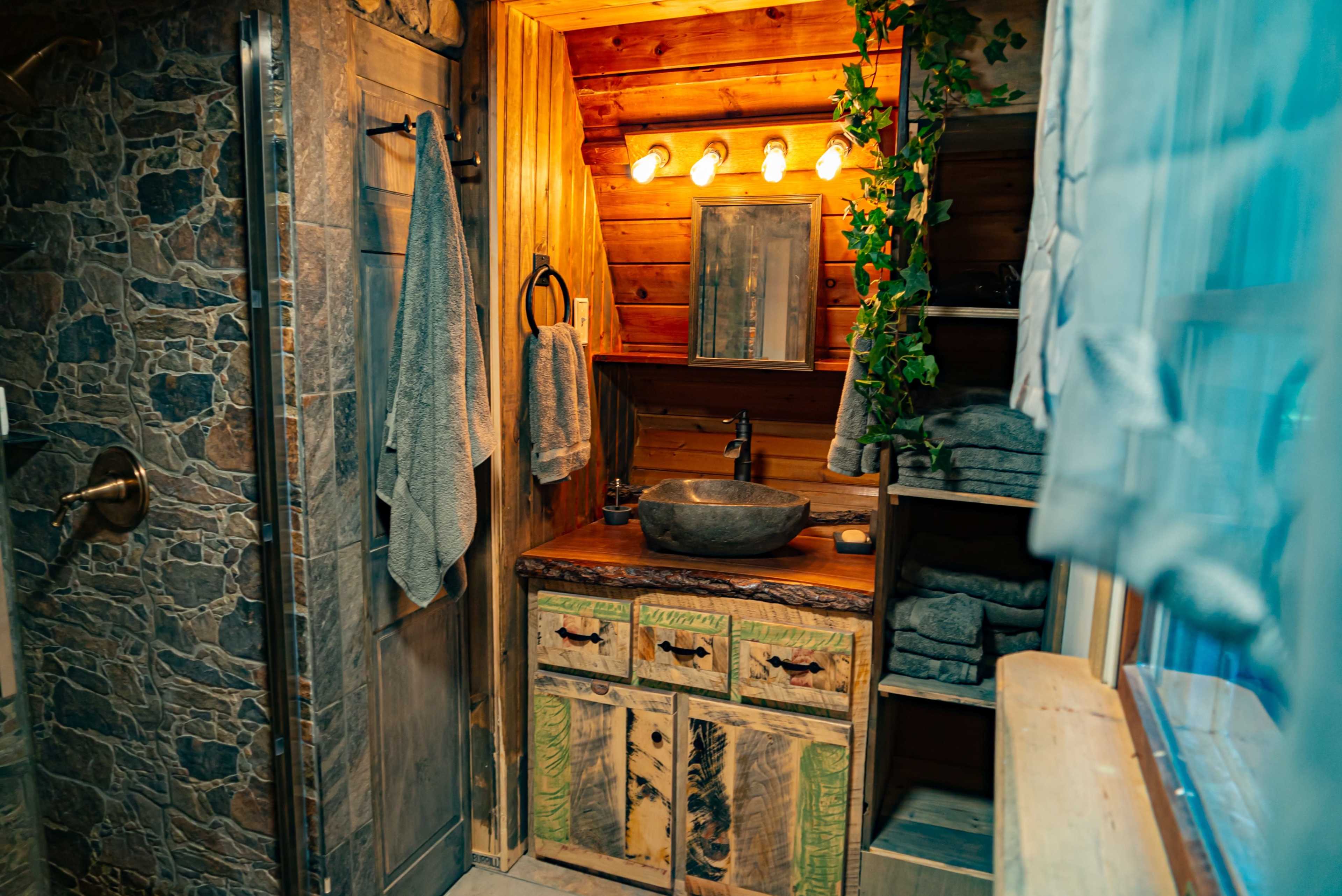 A rustic bathroom with stone walls, wooden accents, a bowl sink on a decorated wooden countertop, and neatly stacked towels.