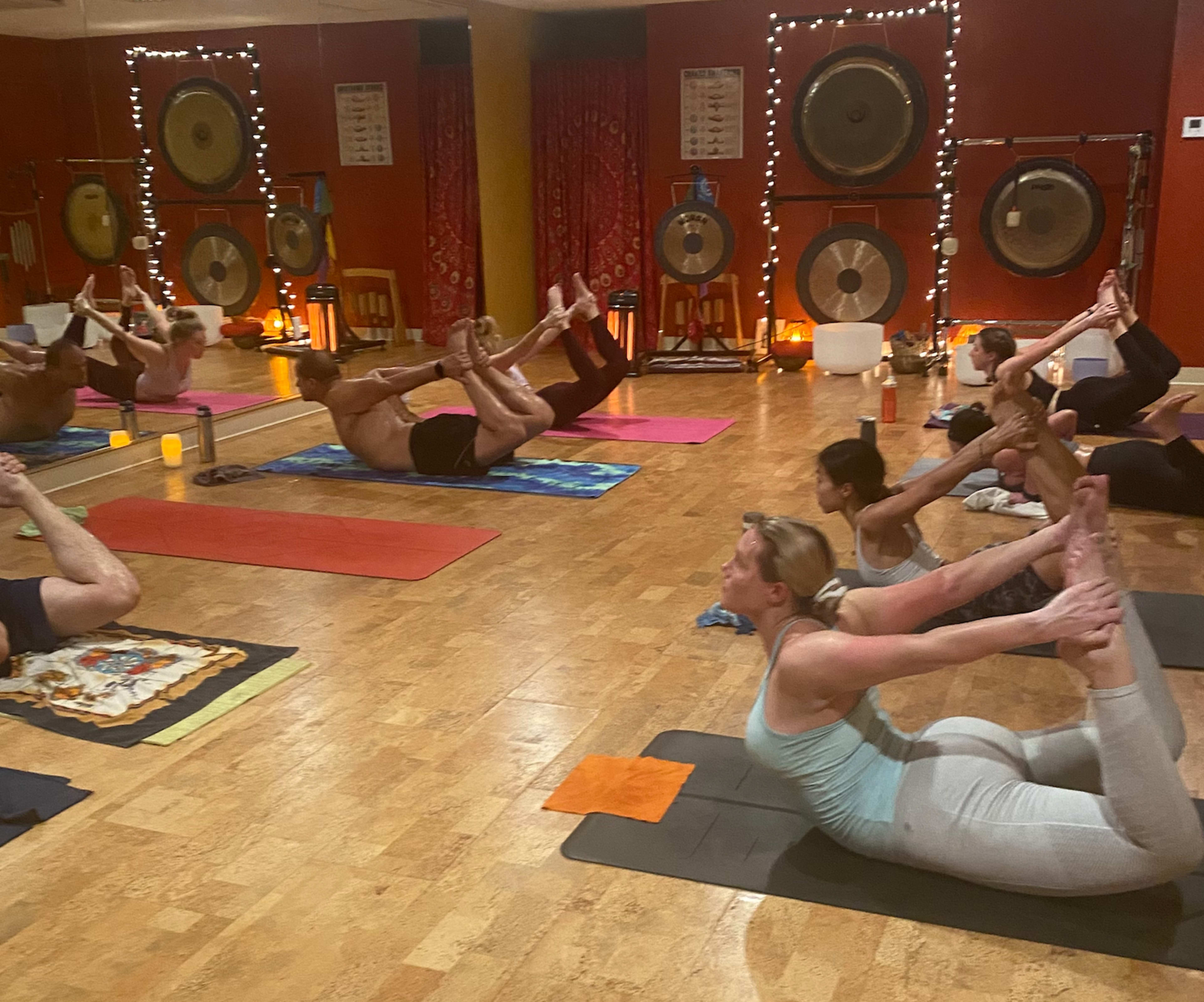 A group of individuals performs yoga poses on mats in a dimly lit studio adorned with gongs and soft lighting.
