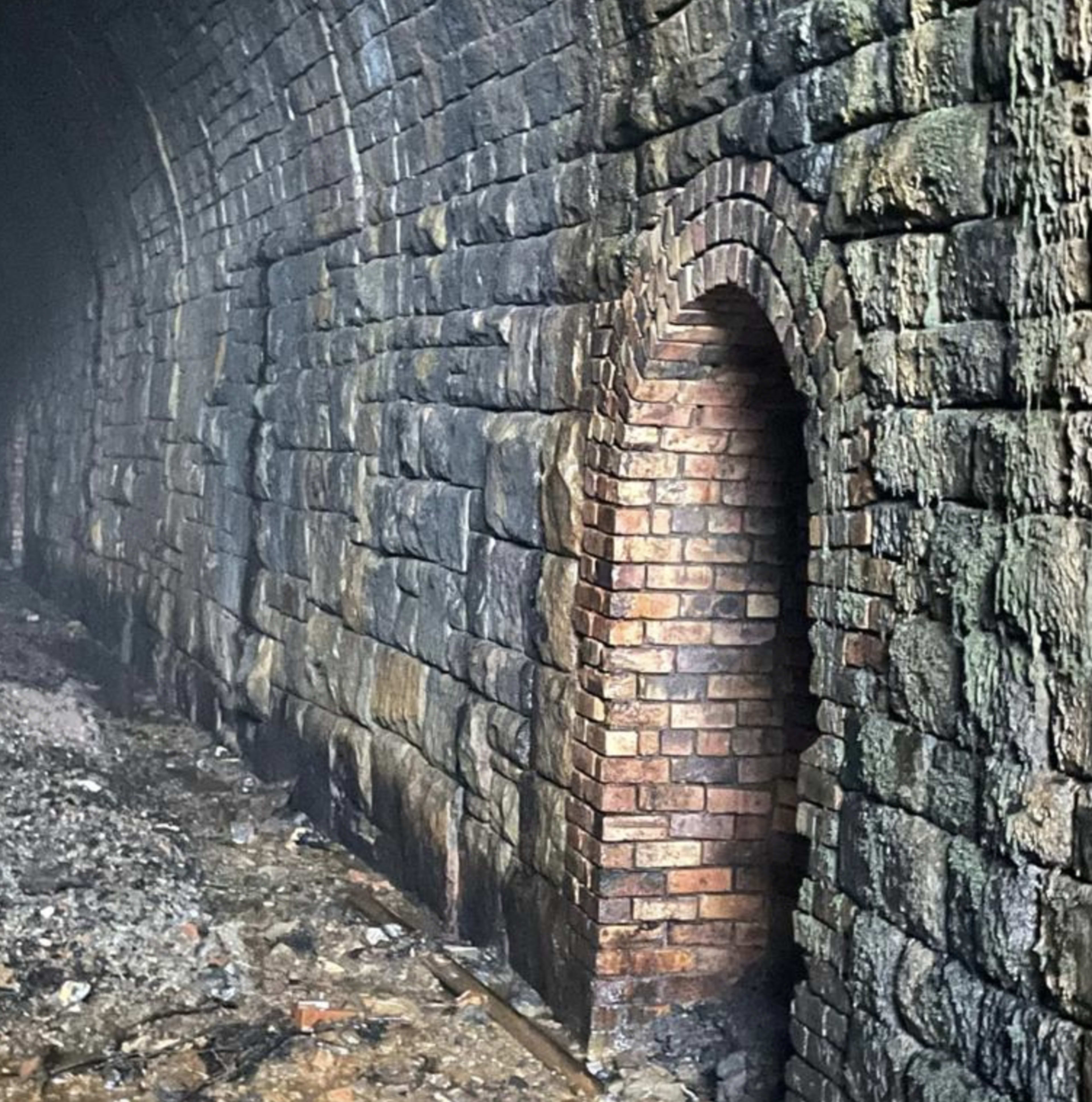 A stone tunnel with a brick archway set into a rough stone wall, partially obscured by dust or debris on the ground.