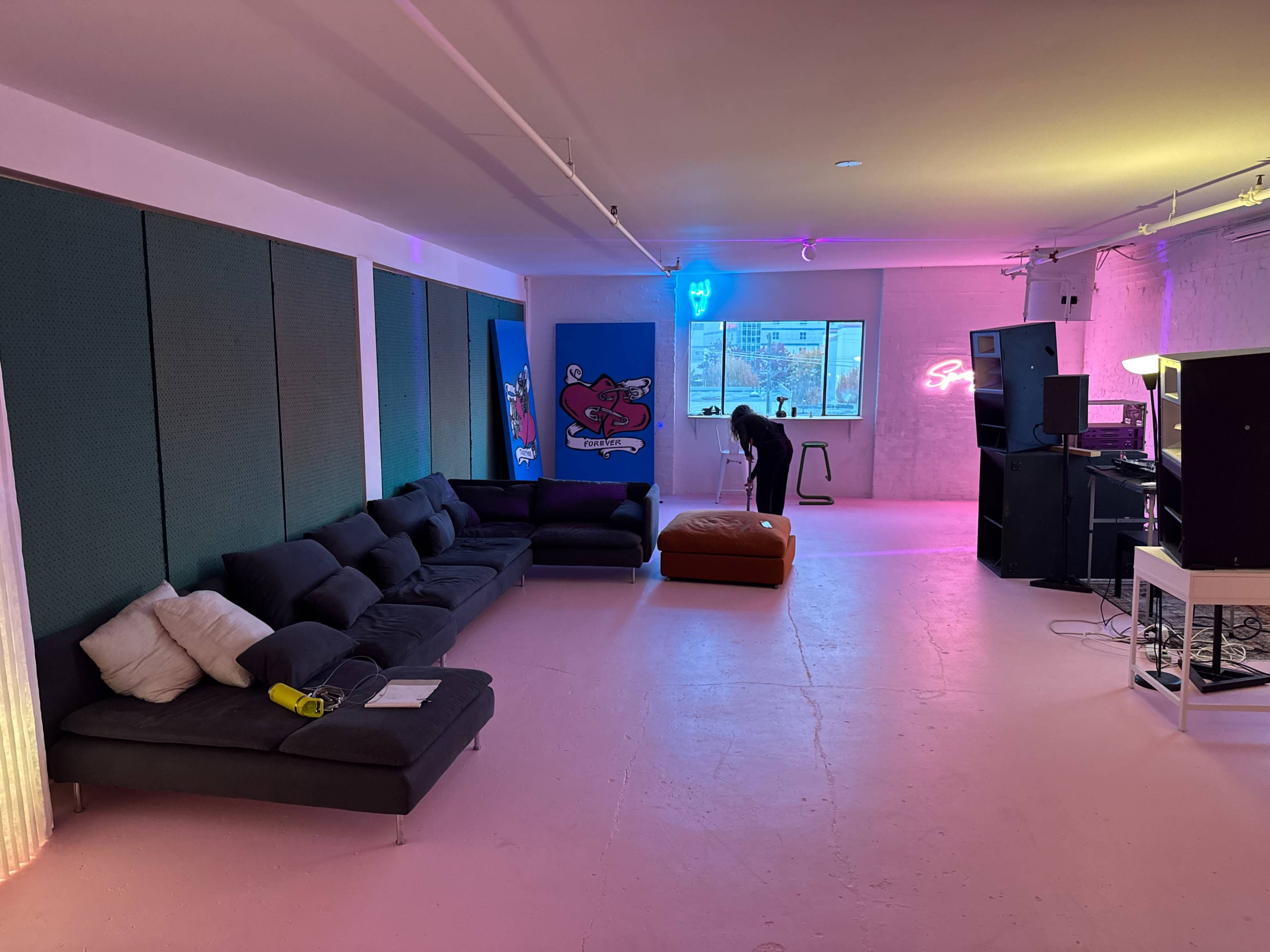 The image shows a spacious, modern room with a large sectional sofa, an orange ottoman, and a music setup in the corner, illuminated by colorful neon lights.