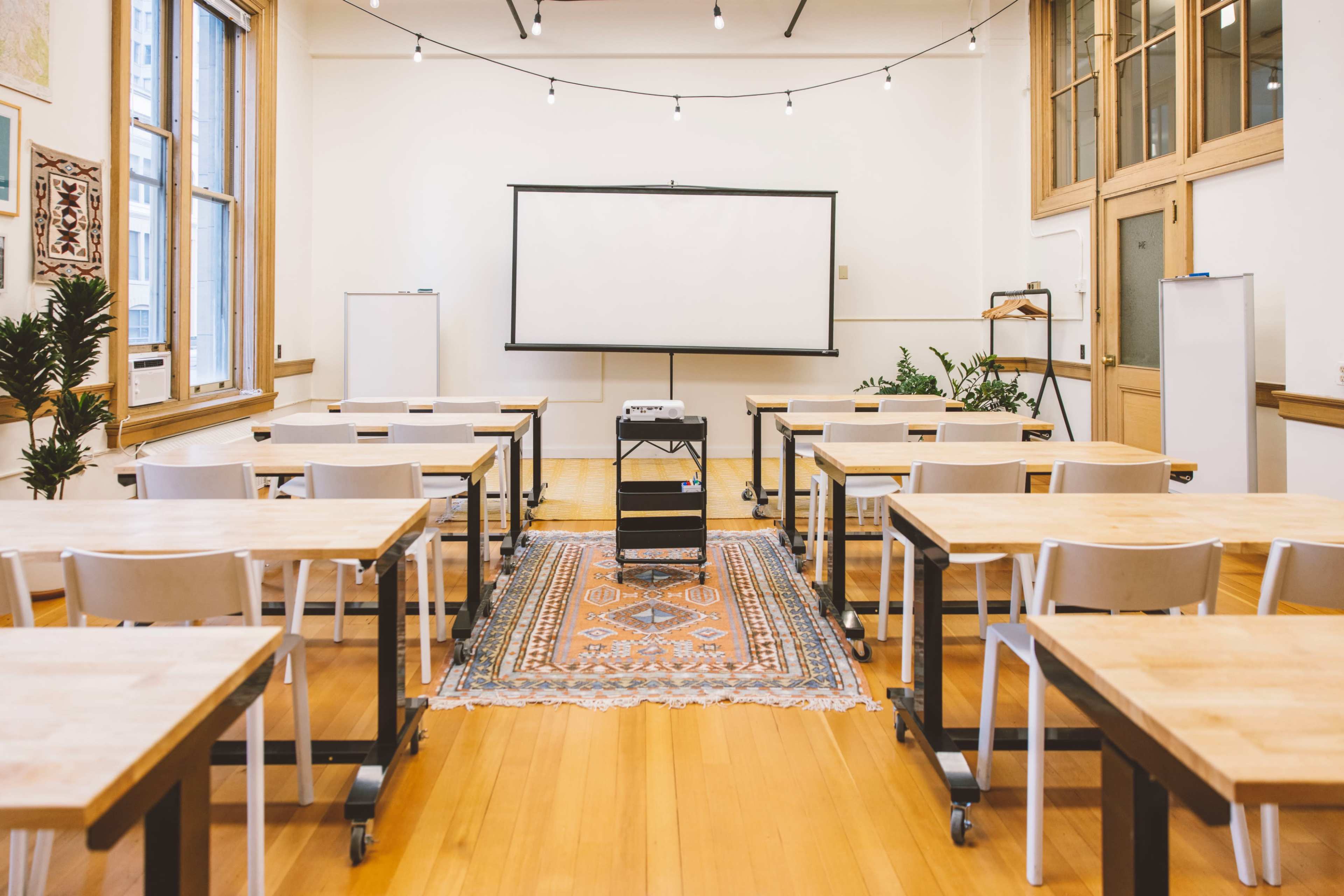 A classroom is set up with rows of tables and chairs facing a blank whiteboard, with a projector and plants arranged in the space.