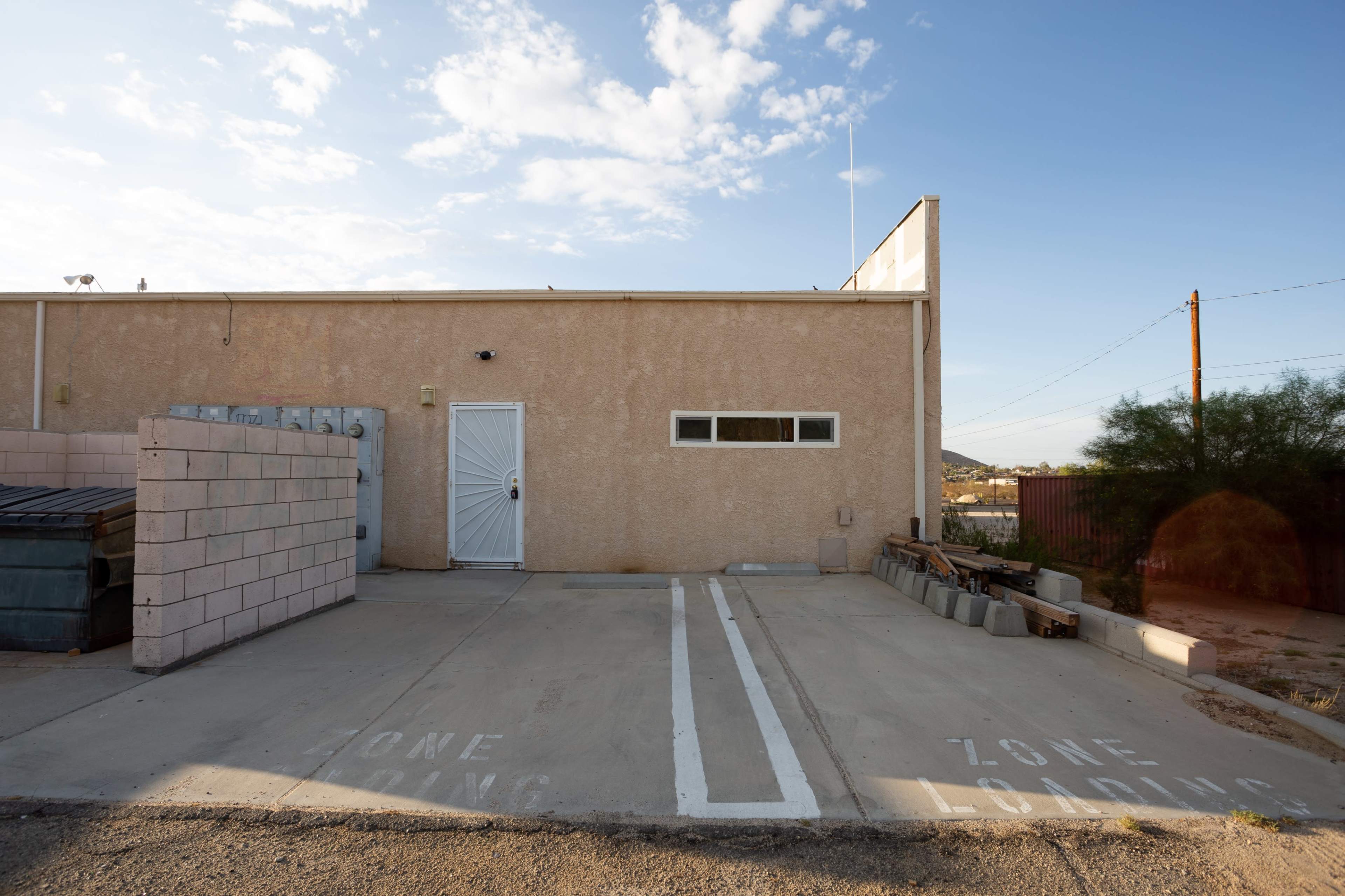 A light-colored building with a loading zone marked on the pavement and a dumpster nearby.