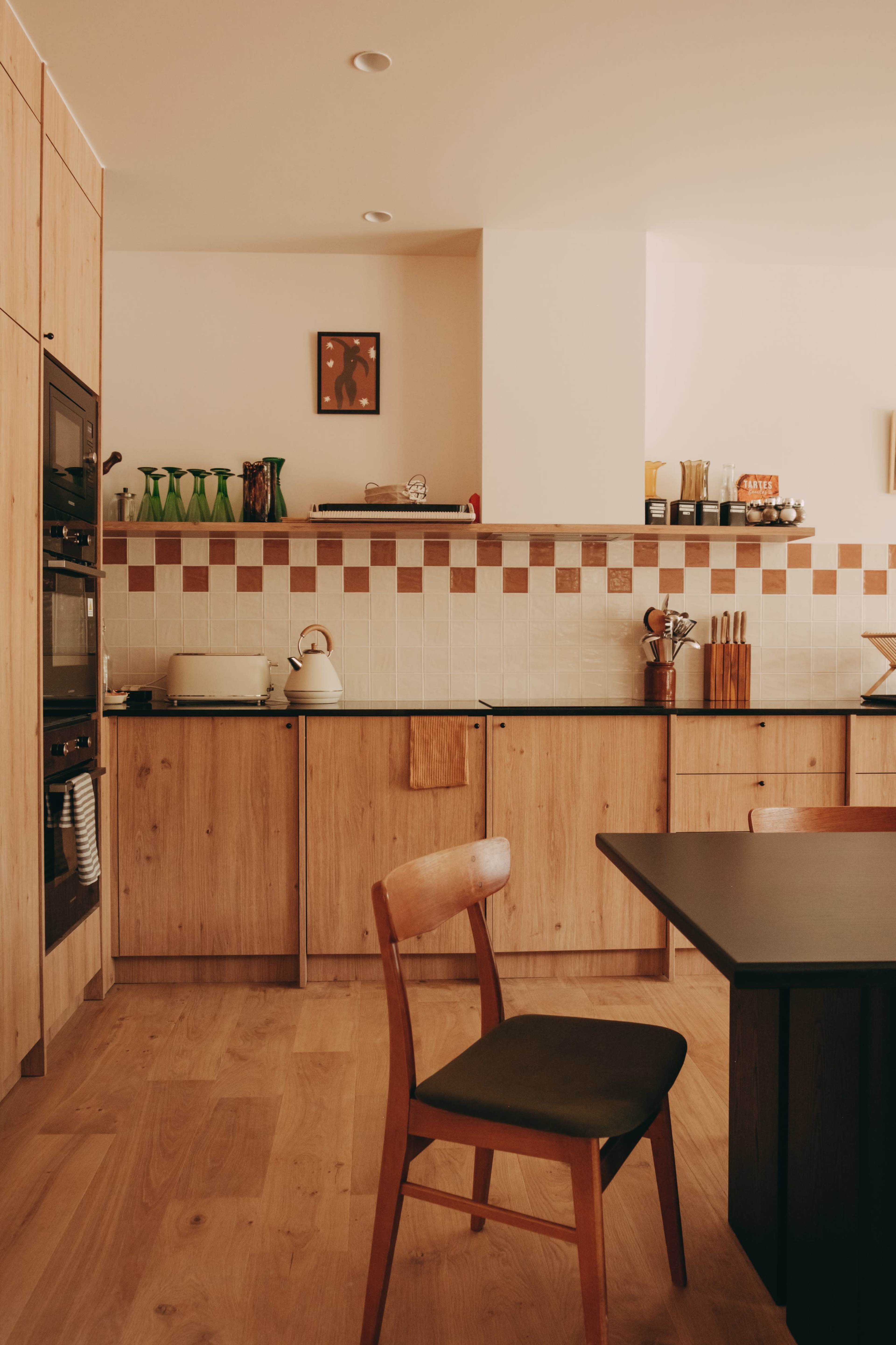 A modern kitchen features wooden cabinetry, a black countertop, and a minimalist dining table with a single chair.