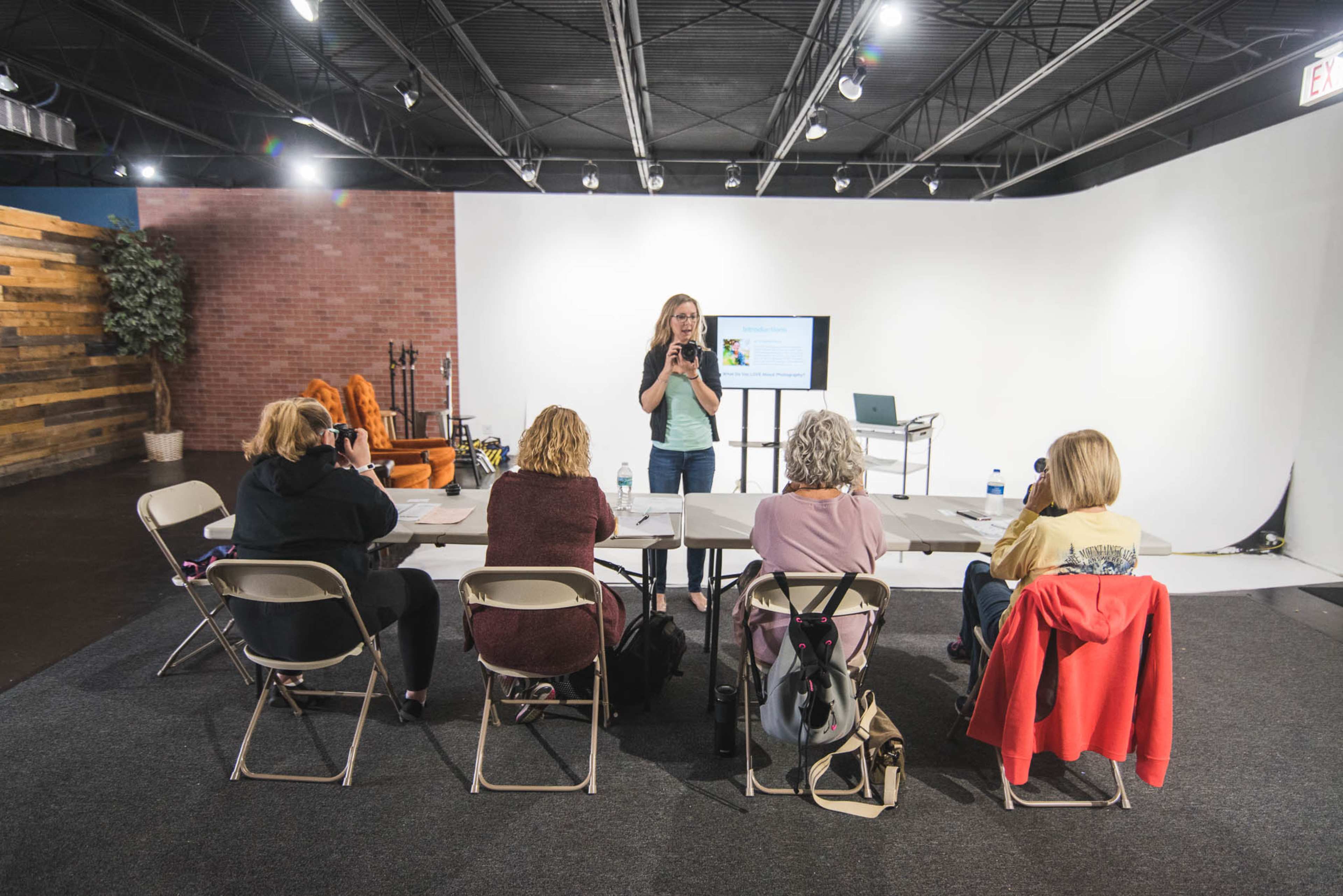 A woman stands at the front of a brightly lit room, presenting to a group of four seated individuals at a table.