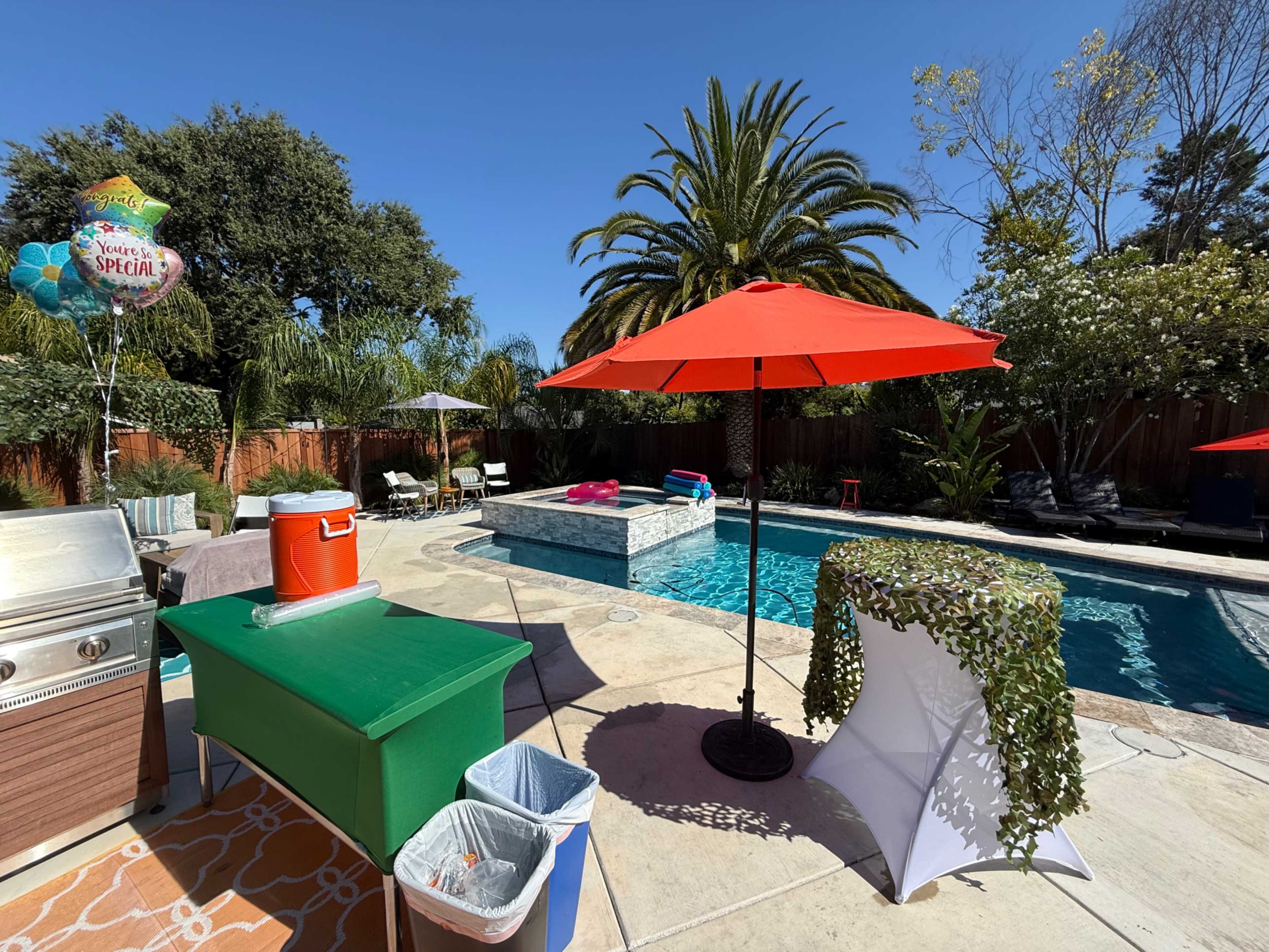 A backyard scene features a swimming pool, various seating areas, a grill, and colorful umbrellas, set against a clear blue sky.