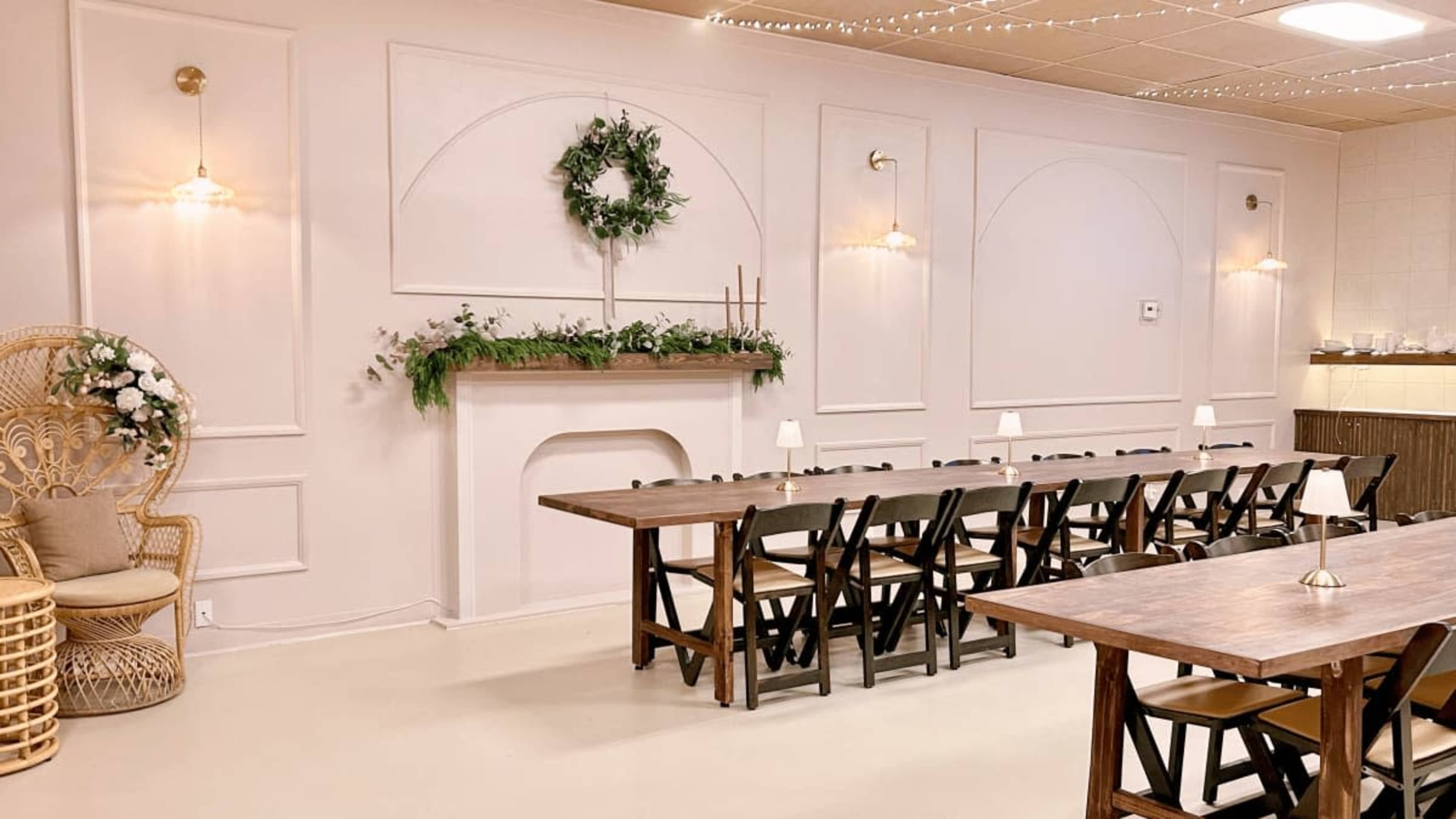 The image shows a spacious, decorated room with a long wooden table, black chairs, and a white mantelpiece adorned with greenery.