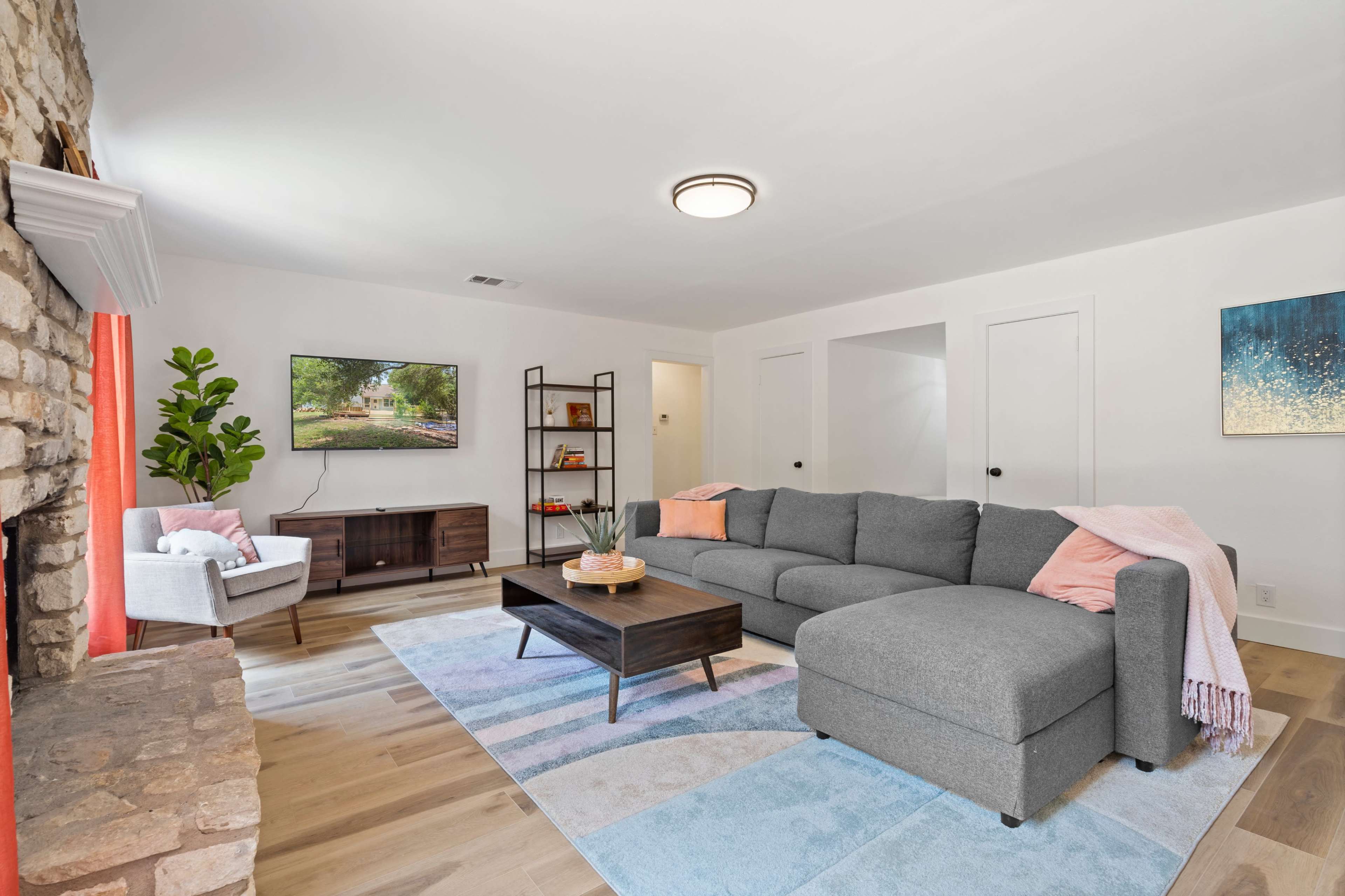 A modern living room features a gray sectional sofa, a wooden coffee table, a flat-screen TV mounted on the wall, and a small armchair beside a stone fireplace.