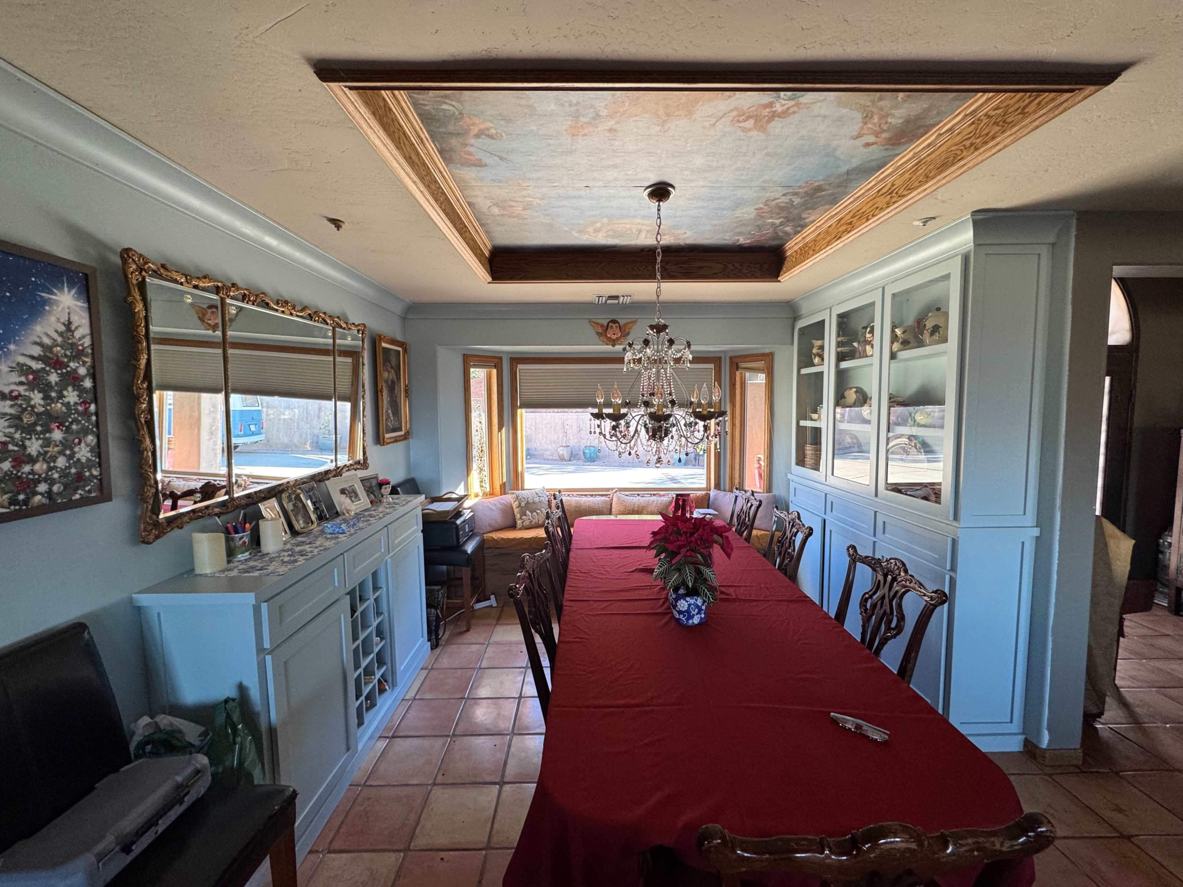 The image shows a dining room with a long table set with a red tablecloth, surrounded by wooden chairs, and featuring a decorative ceiling, a chandelier, and built-in cabinetry.