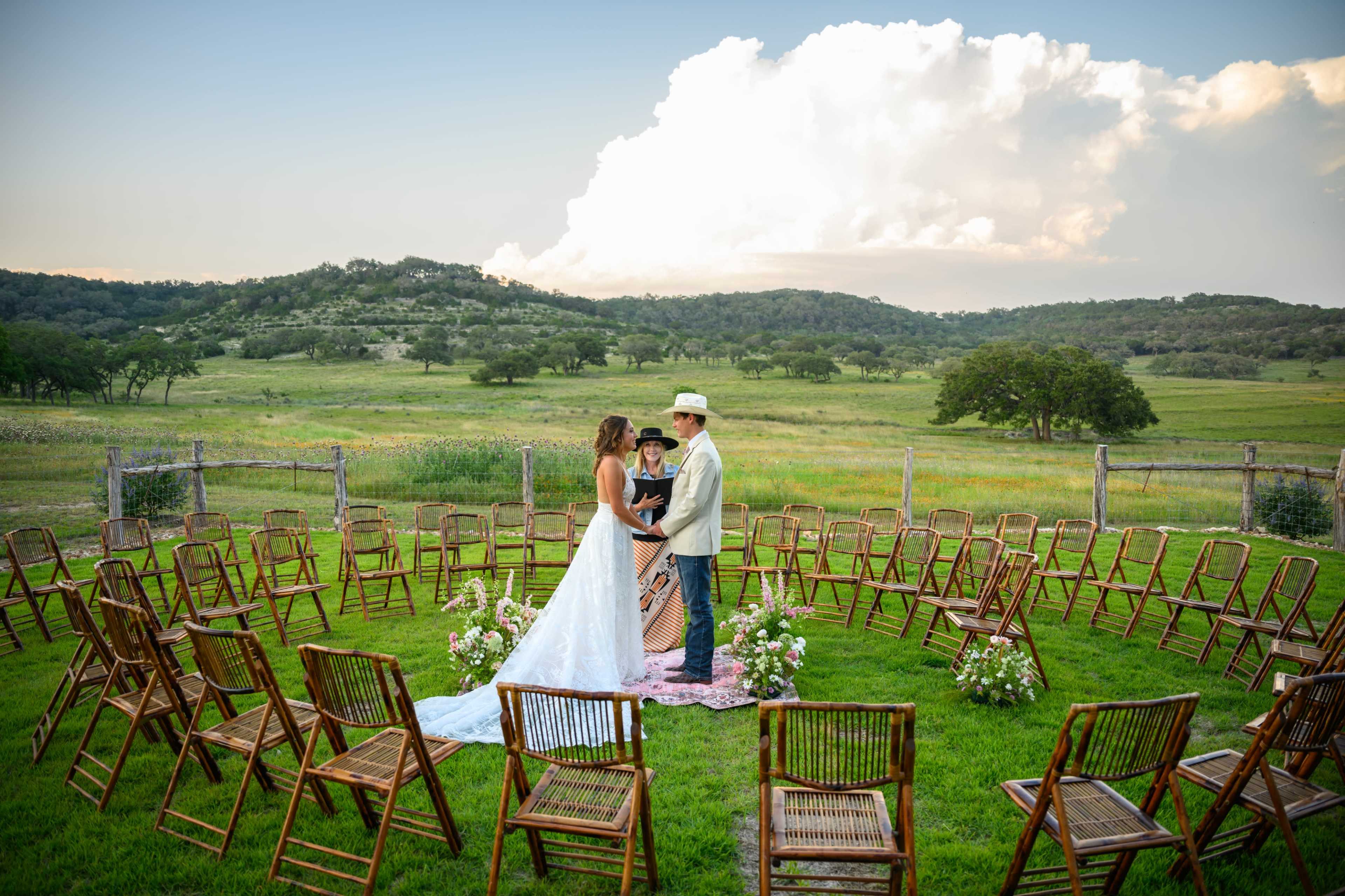 A couple stands in a grassy clearing surrounded by a circle of empty wooden chairs, with rolling hills and clouds in the background.