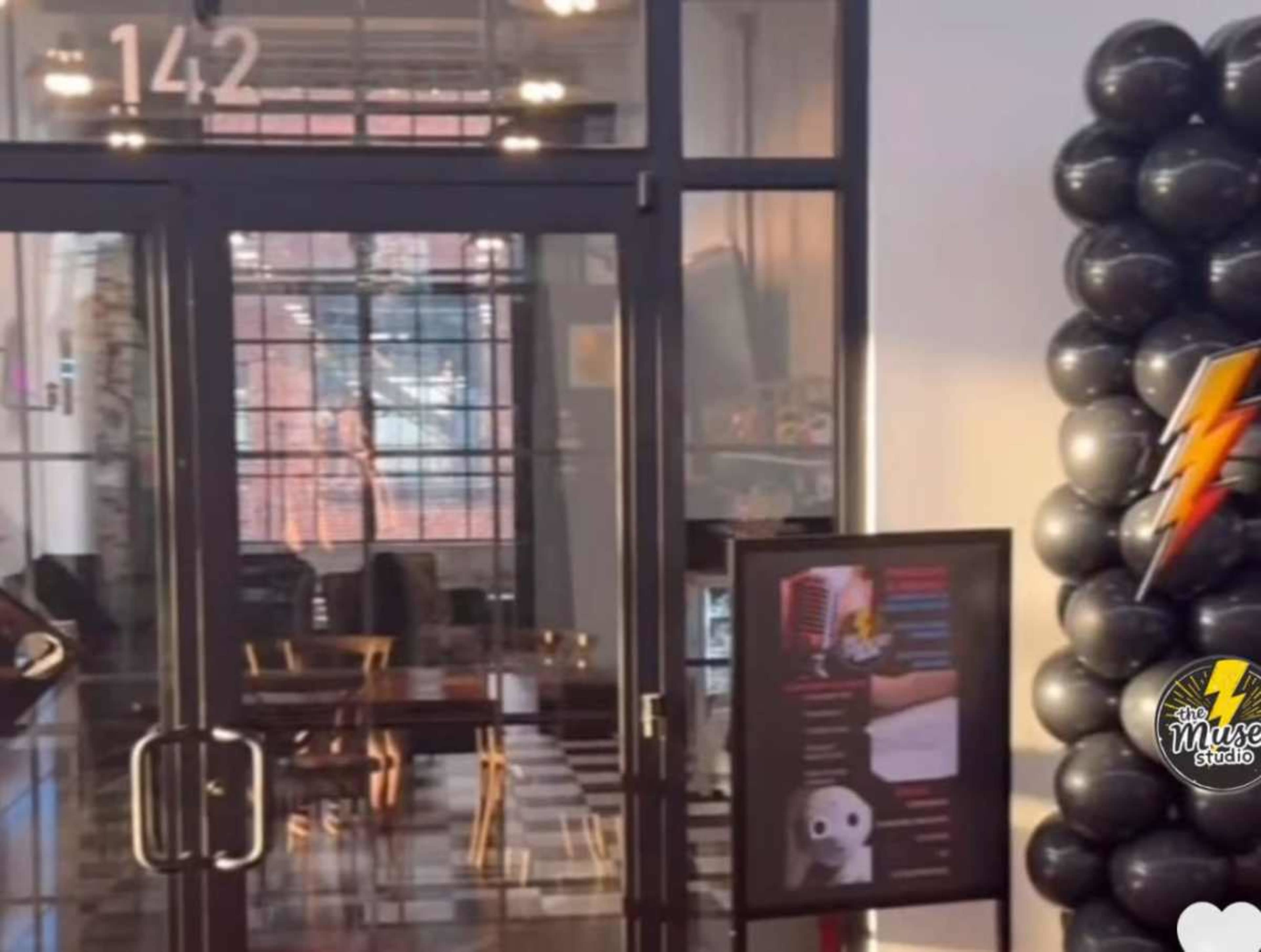 The image shows the entrance of a studio with large glass doors, a welcoming black balloon arch to the right, and a sign displaying information in front.