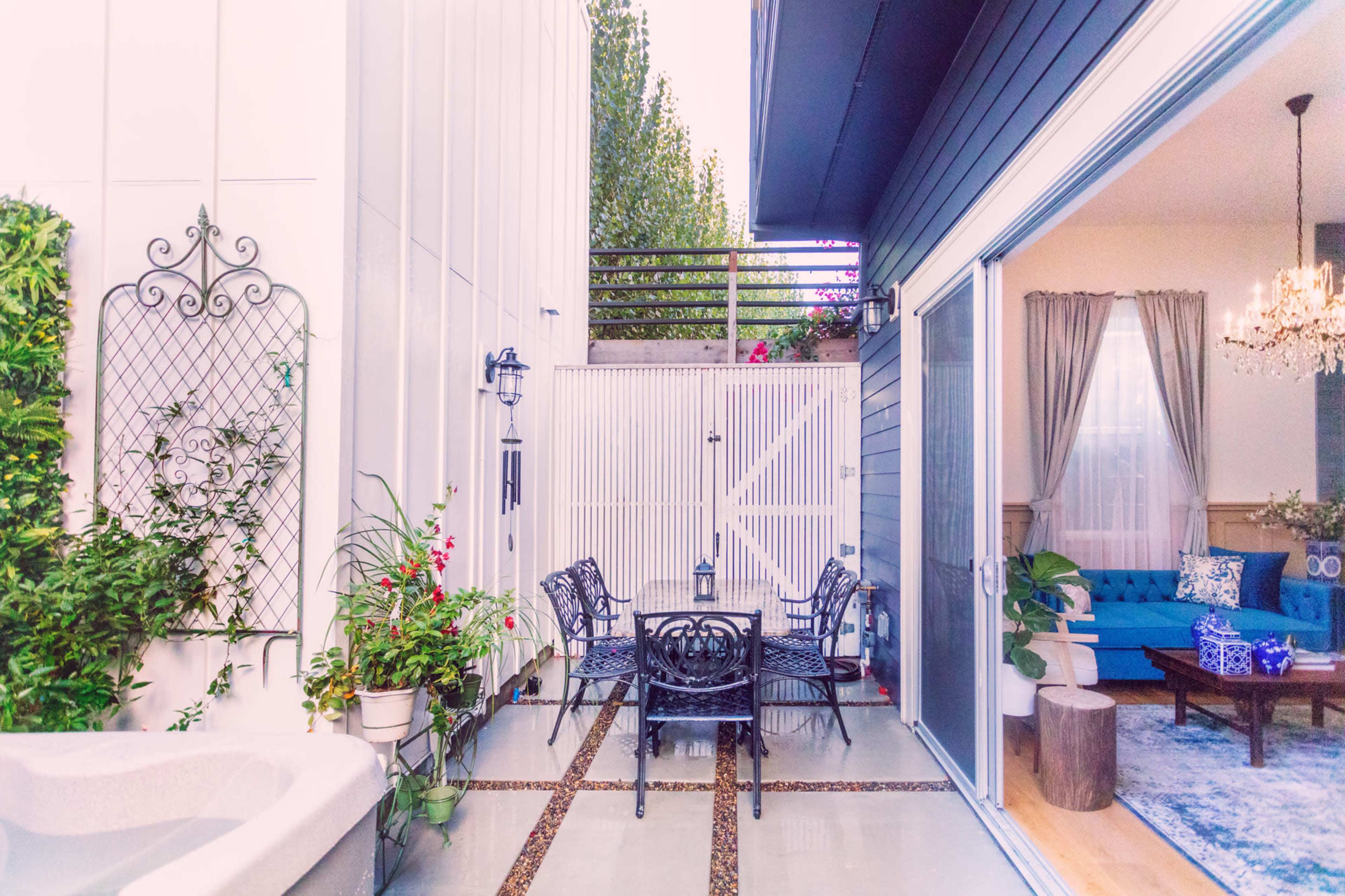 A patio area features a dining table with four black metal chairs, surrounded by plants, next to a white wooden fence, and a blue sofa is visible through a large glass door.
