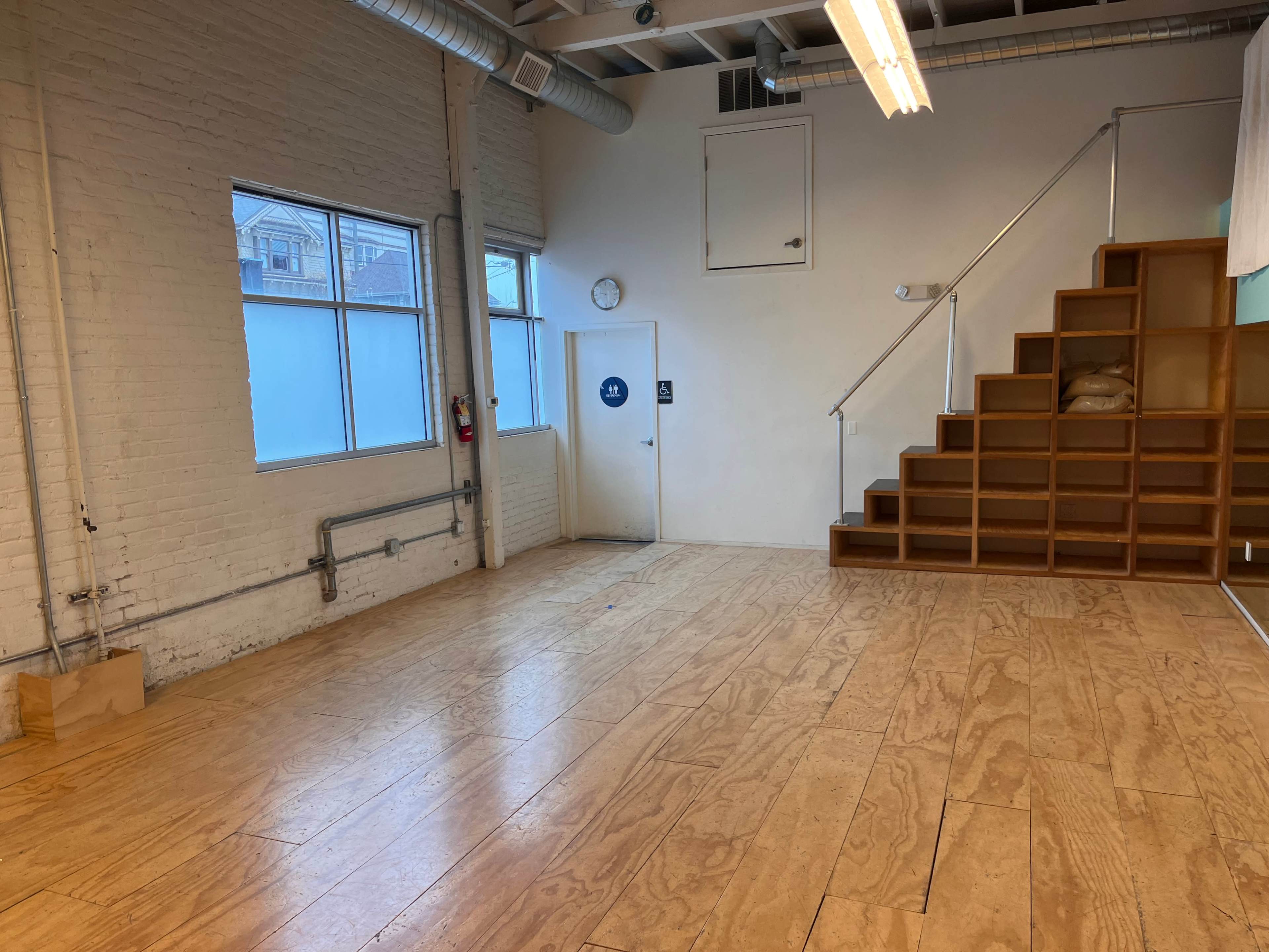 A spacious room with wooden flooring, a set of stairs on the right leading to a loft area, and large windows on the left.