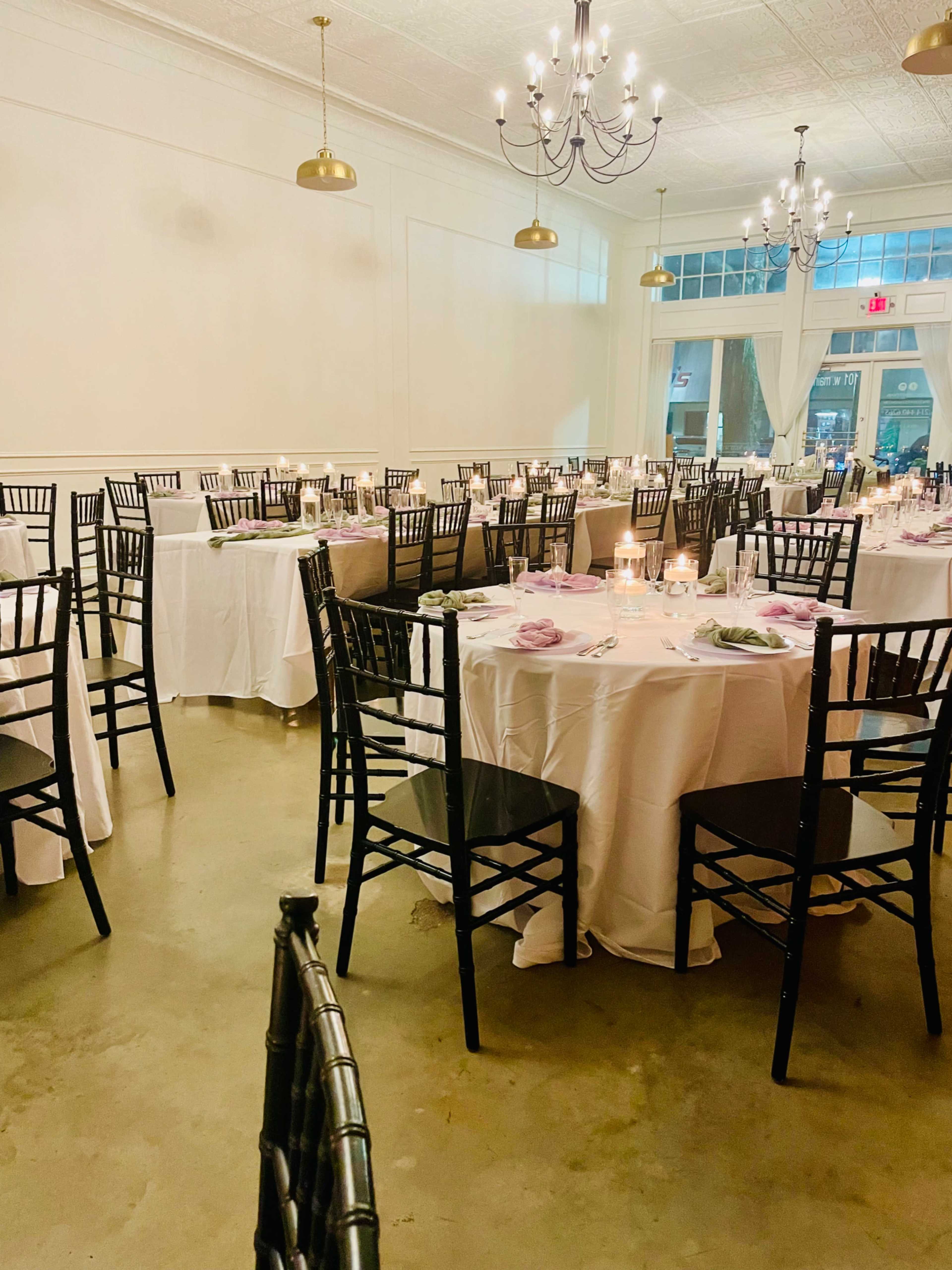 The image shows a restaurant setting with neatly arranged round tables covered in white tablecloths and black chairs, illuminated by soft lighting from chandeliers.