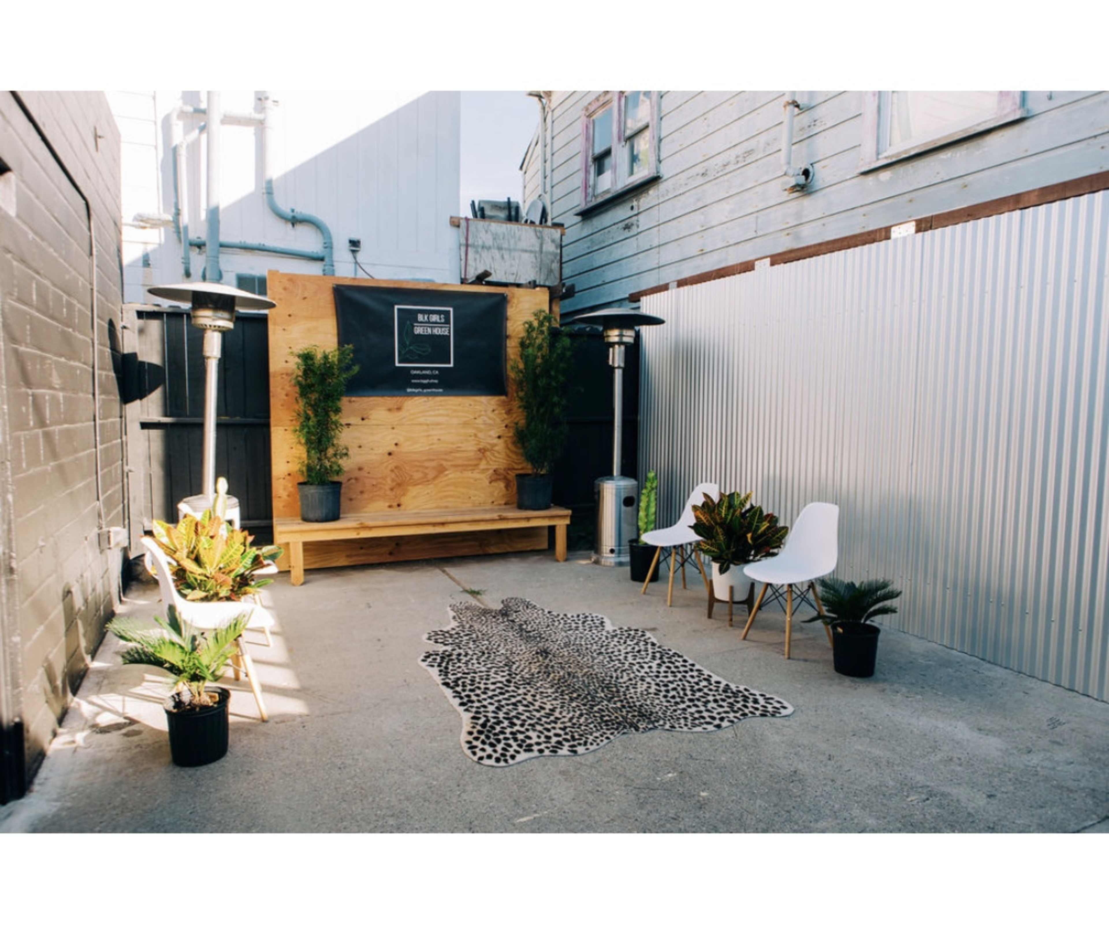 A small outdoor seating area with two white chairs, a wooden bench, potted plants, and a faux animal skin rug on a concrete floor, surrounded by walls of wood and metal.