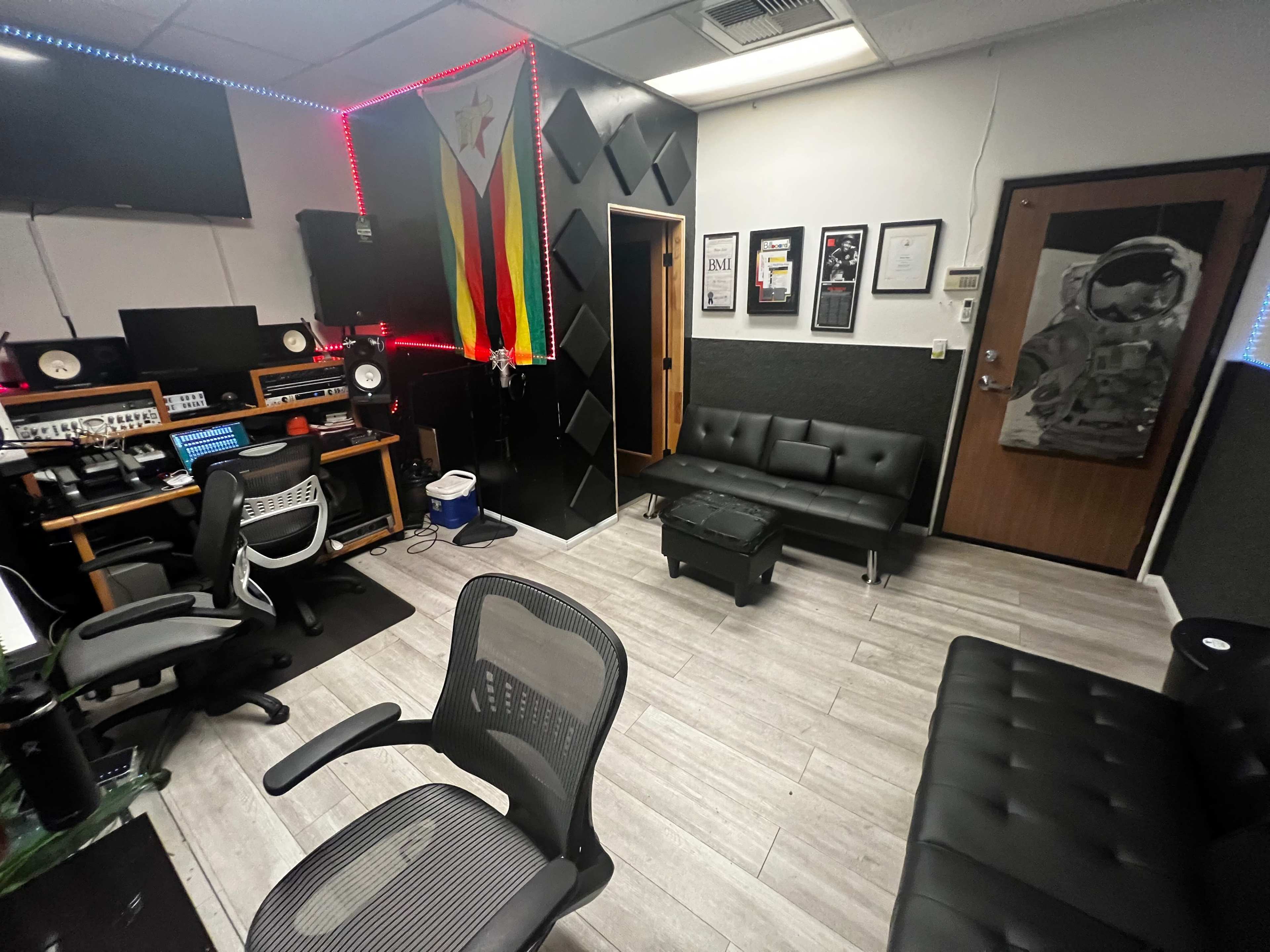 The image shows a recording studio with a sound setup, black chairs, a colorful flag on the wall, and various framed certificates.