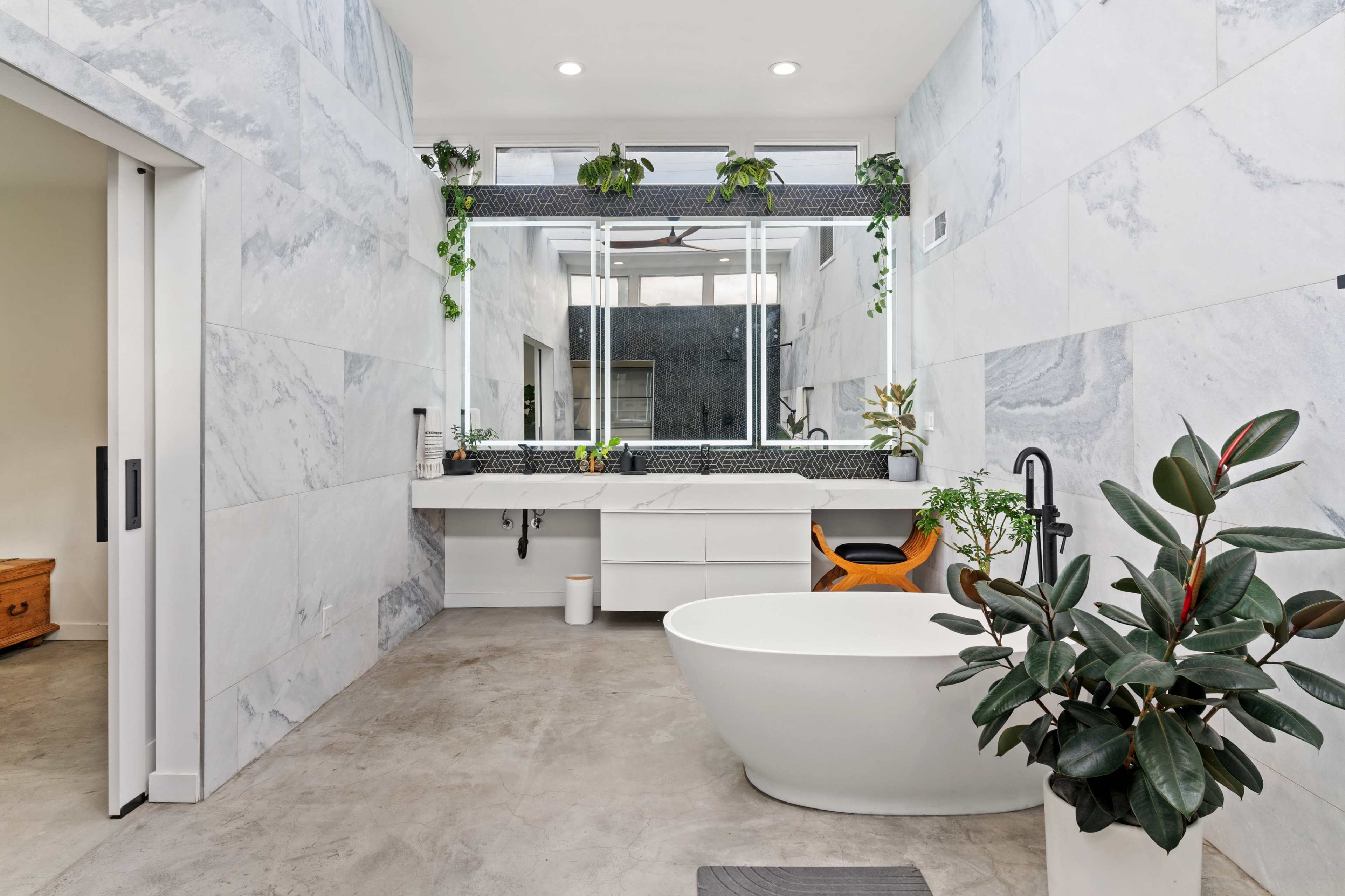 The bathroom features marble walls, a freestanding bathtub, a large mirror, and various indoor plants.