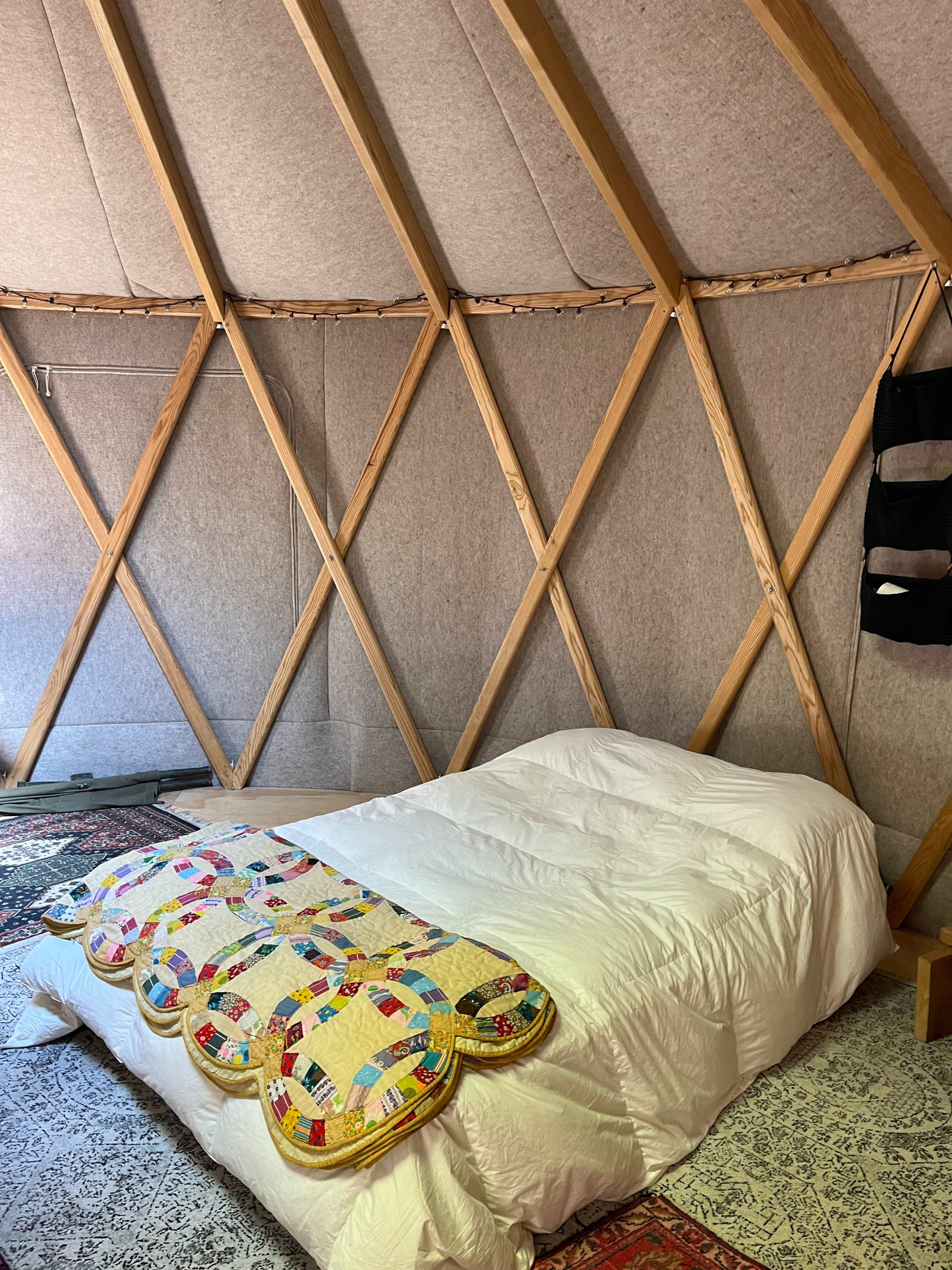 A cozy interior of a yurt with a bed covered by a colorful quilt and wooden support beams lining the walls.