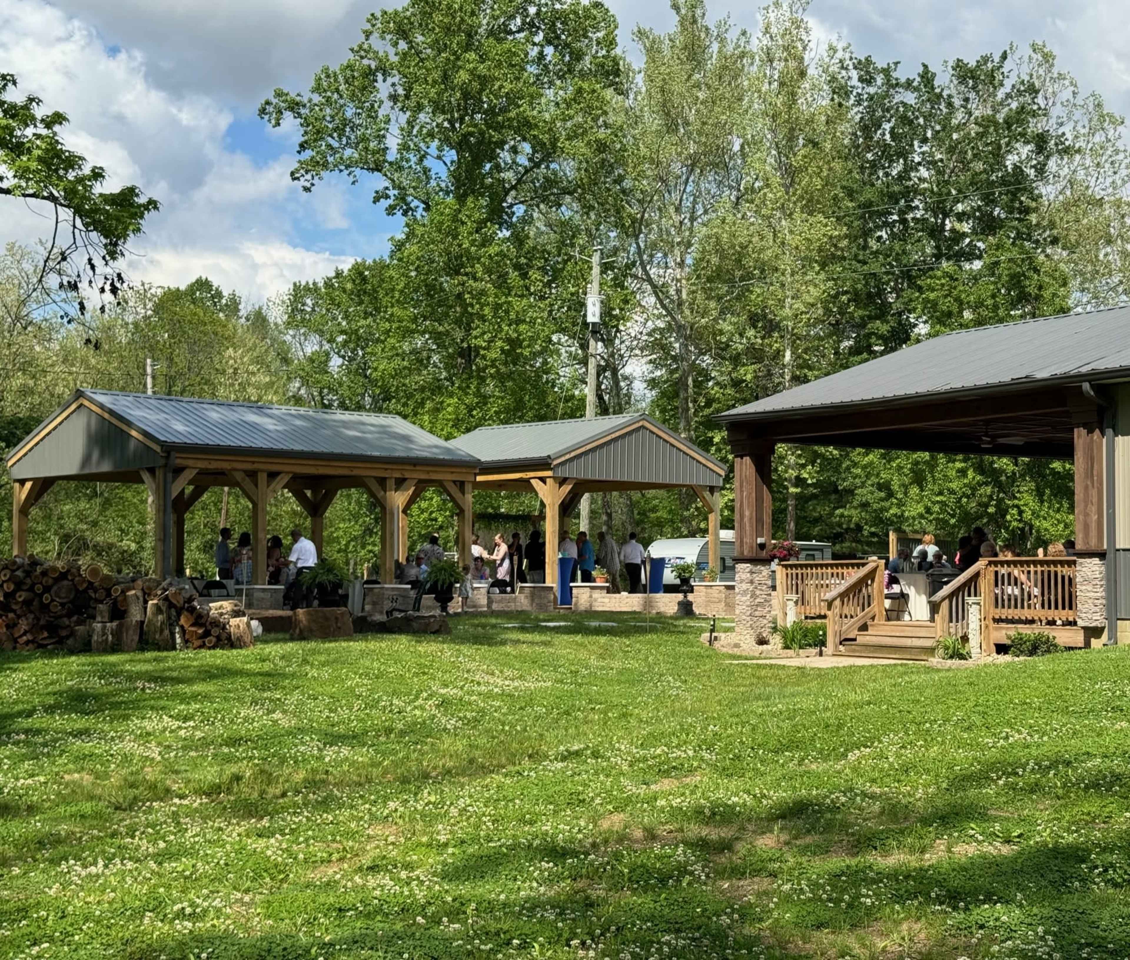 A gathering takes place under wooden pavilions in a grassy area surrounded by trees.