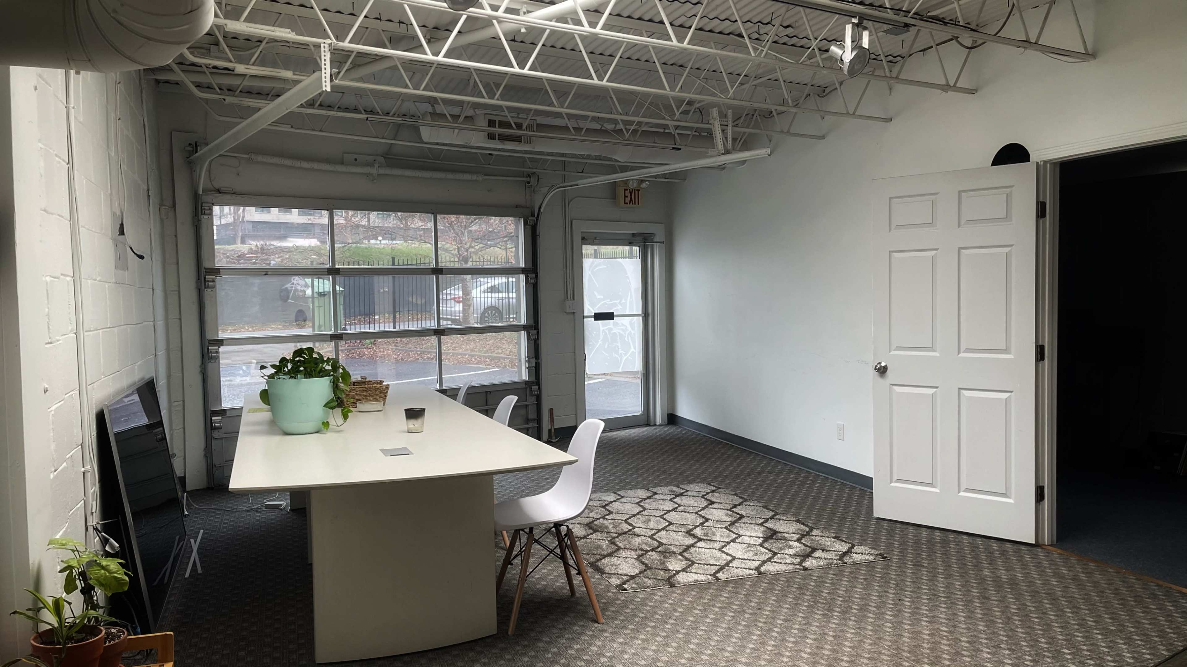 A simple interior space with a large white table, chairs, plants, and windows, along with a door leading outside.