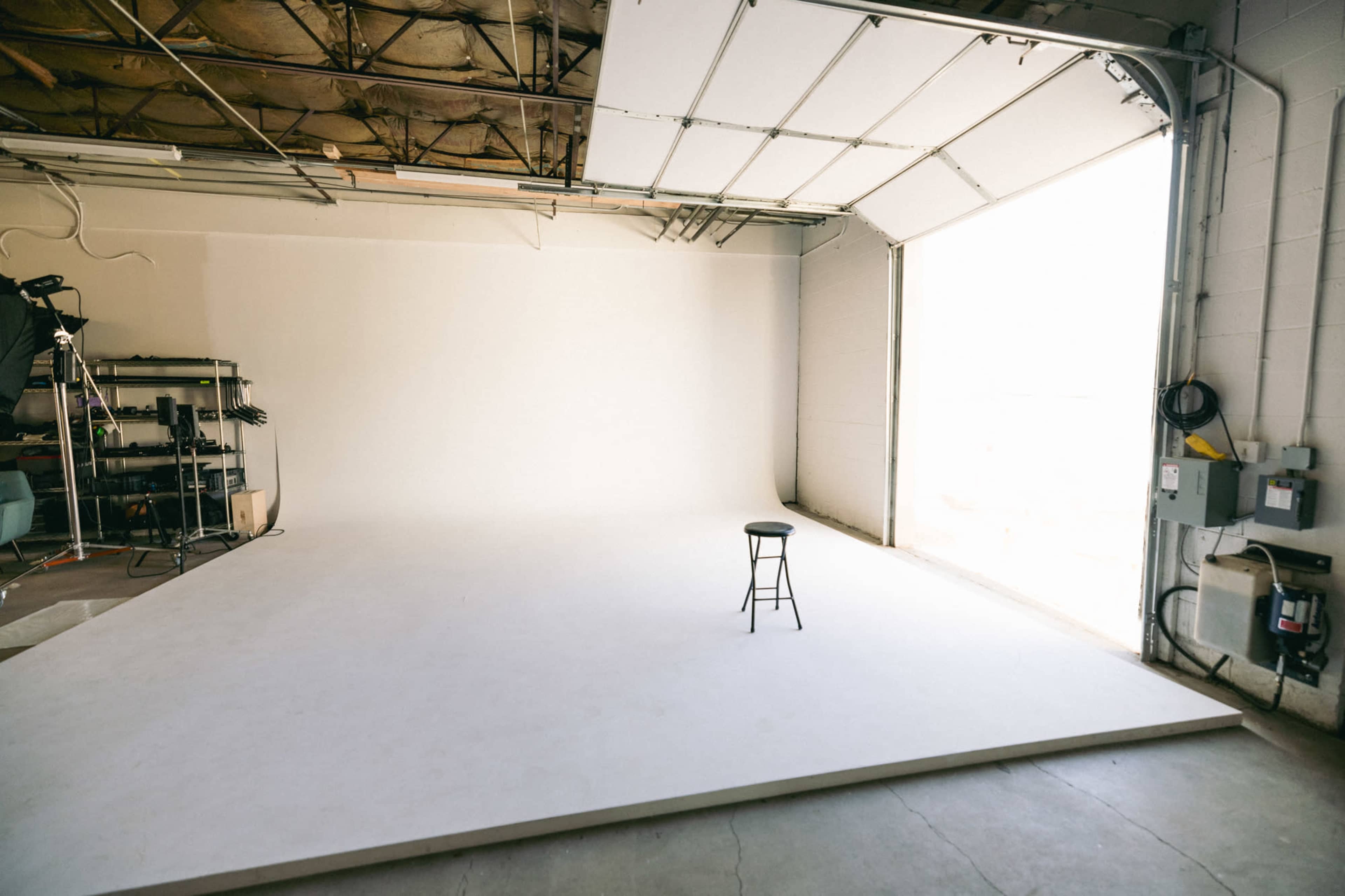A minimalistic photo studio features a single black stool on a white backdrop, illuminated by natural light from an open garage door.