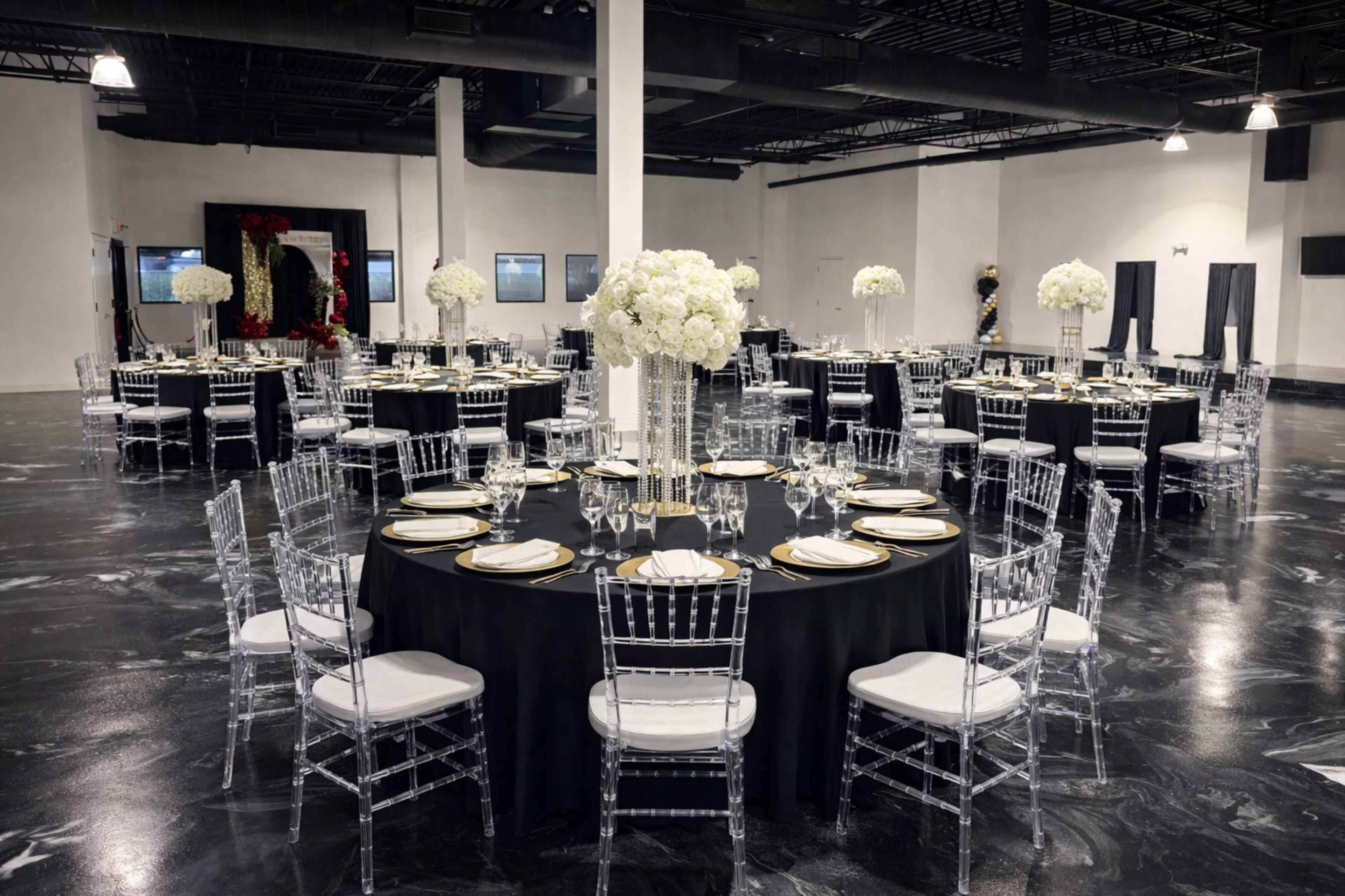 The image shows an elegantly arranged banquet hall with round tables covered in black tablecloths, each featuring tall floral centerpieces and surrounded by clear acrylic chairs.