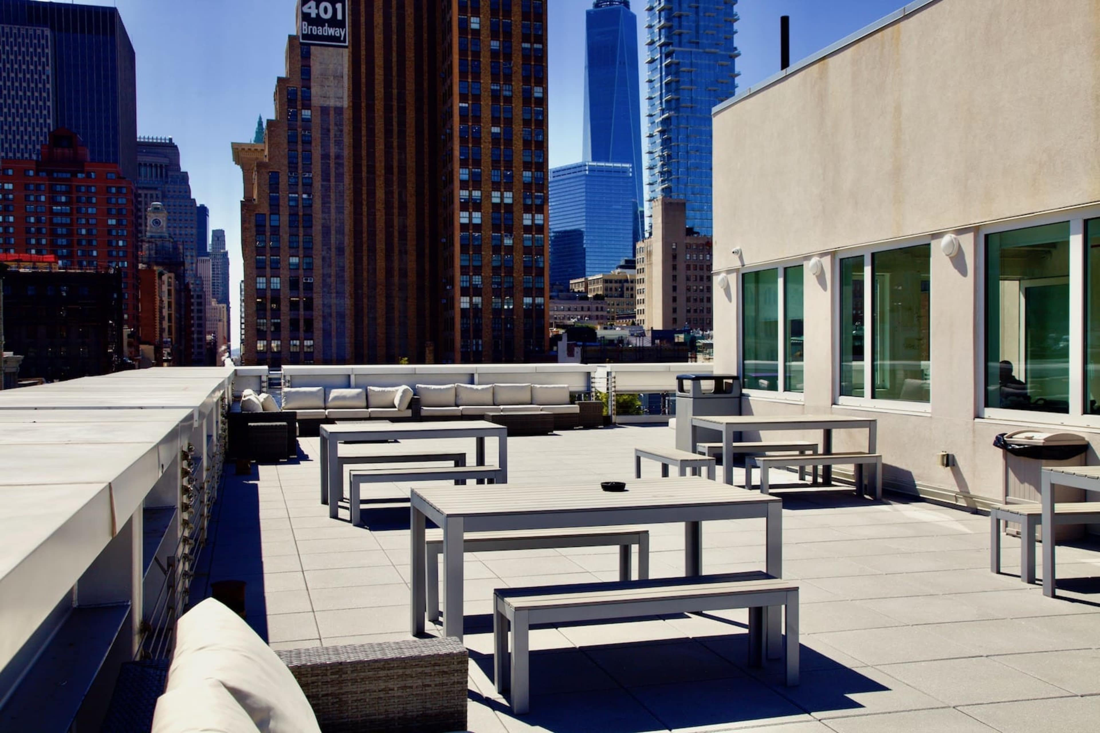 The image shows a rooftop terrace with gray tables, benches, and a clear view of tall buildings in the background.