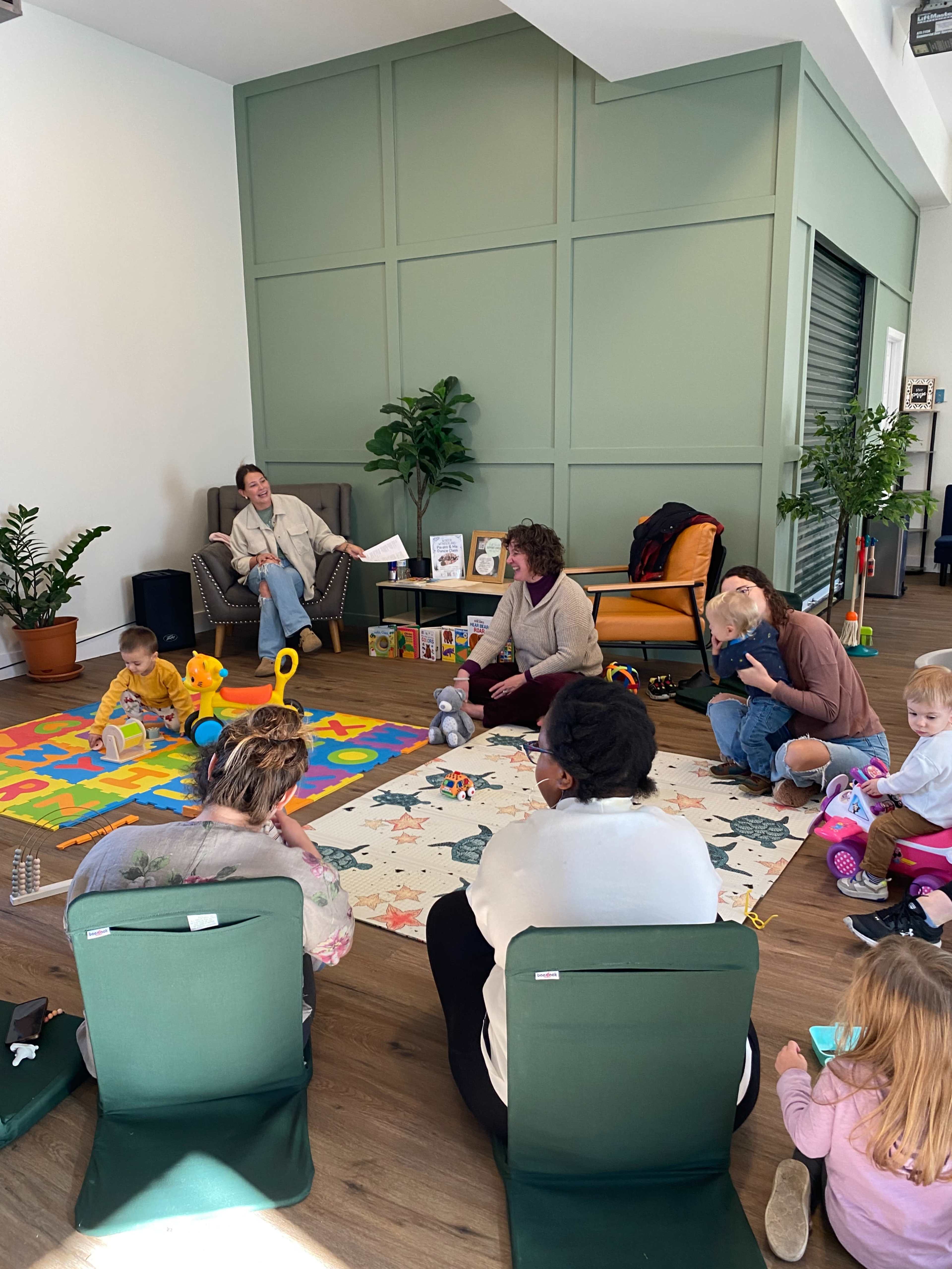 A group of adults and children are seated on the floor of a bright, modern room, surrounded by colorful play mats and toys, while another adult sits in a chair and supervises.