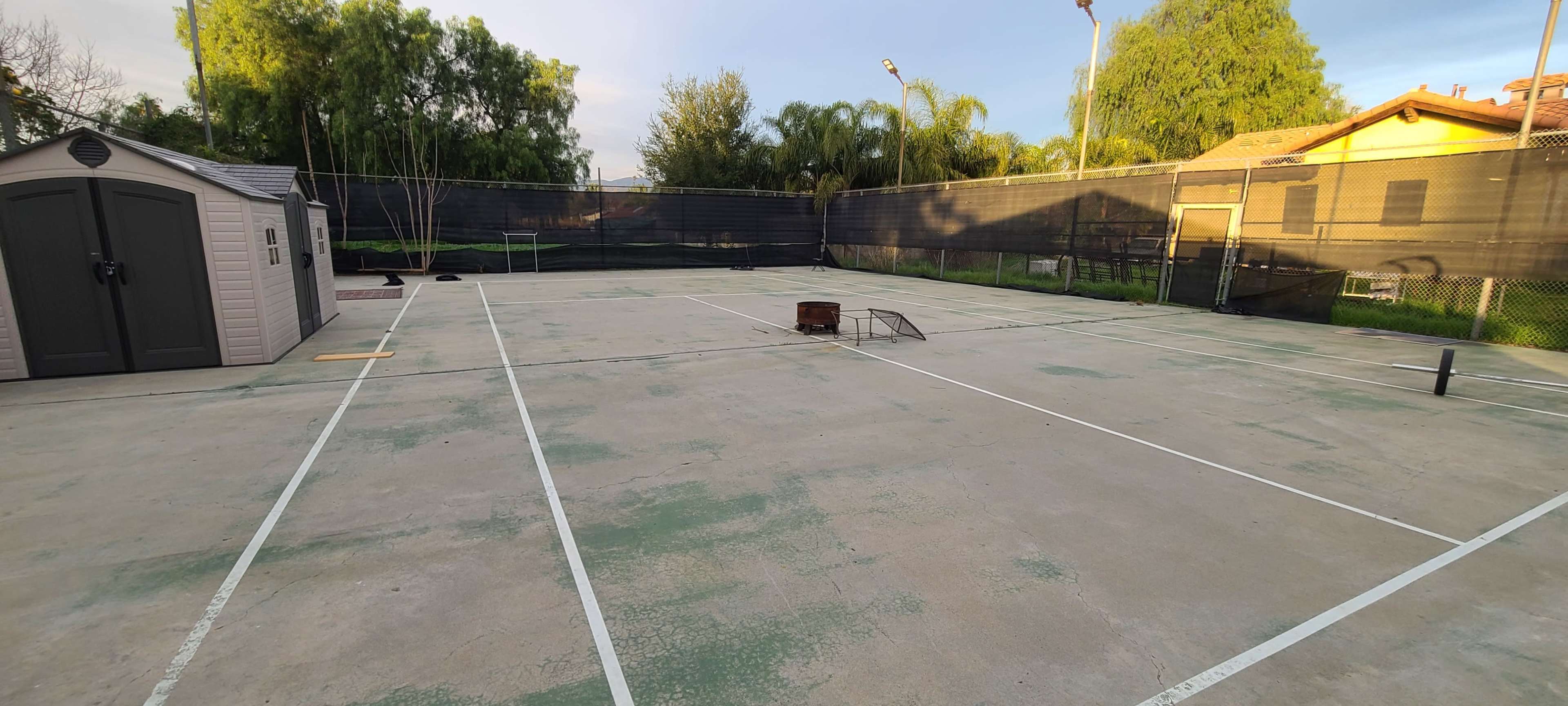 A tennis court with a storage shed in one corner and a few trees in the background under clear skies.