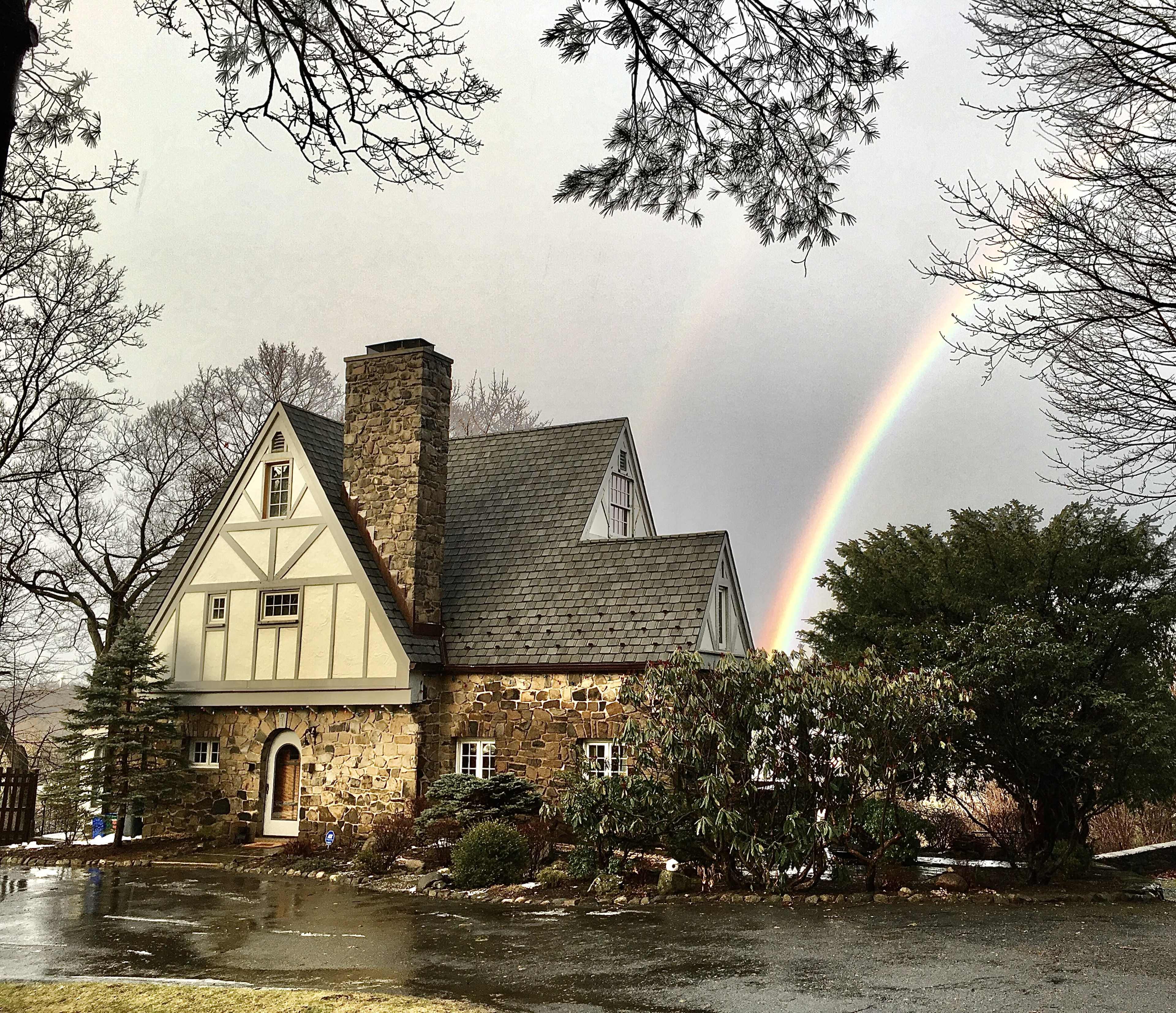 A quaint stone and timber-framed house sits under a gray sky, with a rainbow arcing above it and trees surrounding the property.