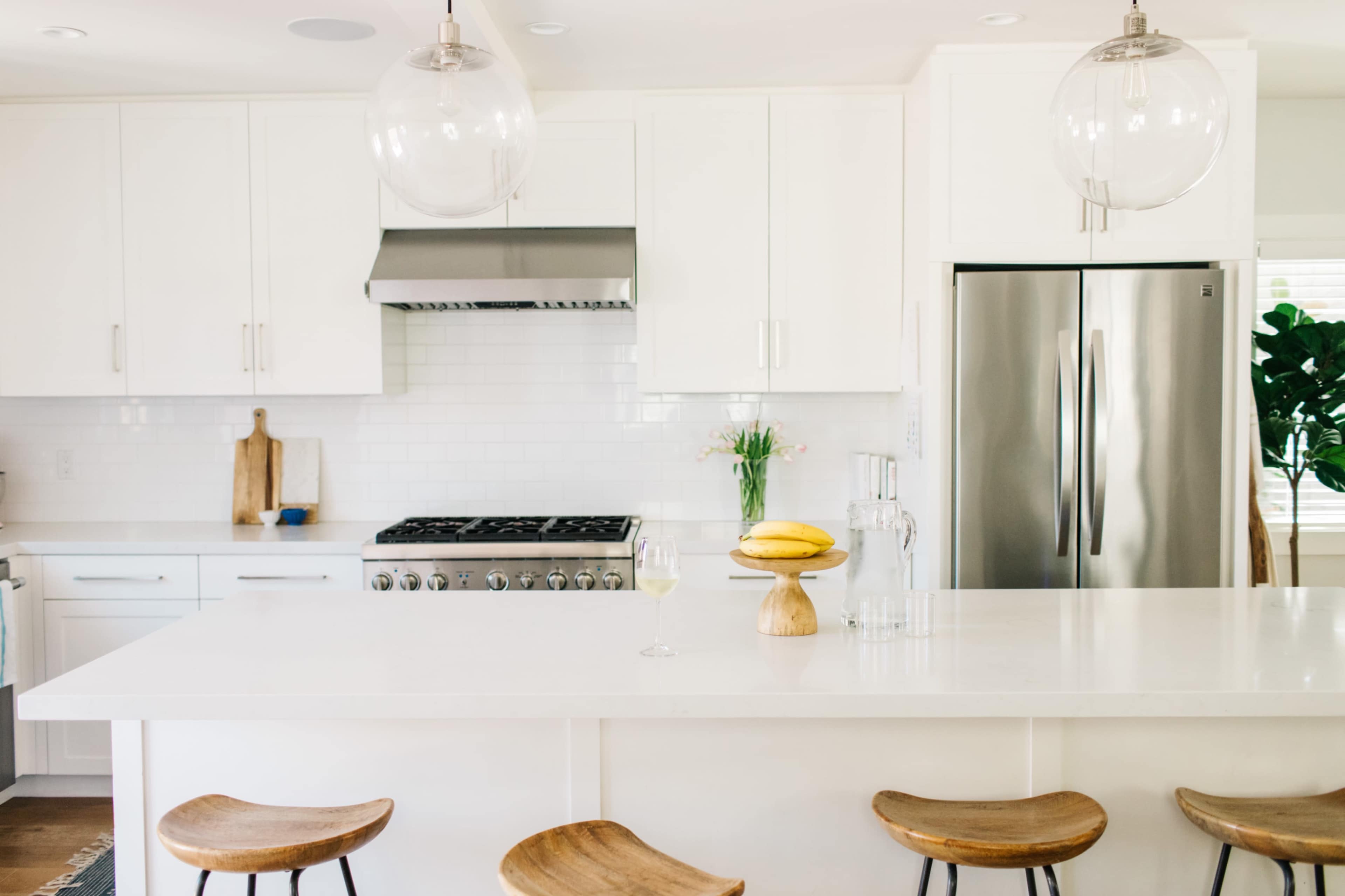 A modern kitchen with white cabinetry, stainless steel appliances, and a central island topped with a wooden display of bananas and a glass.