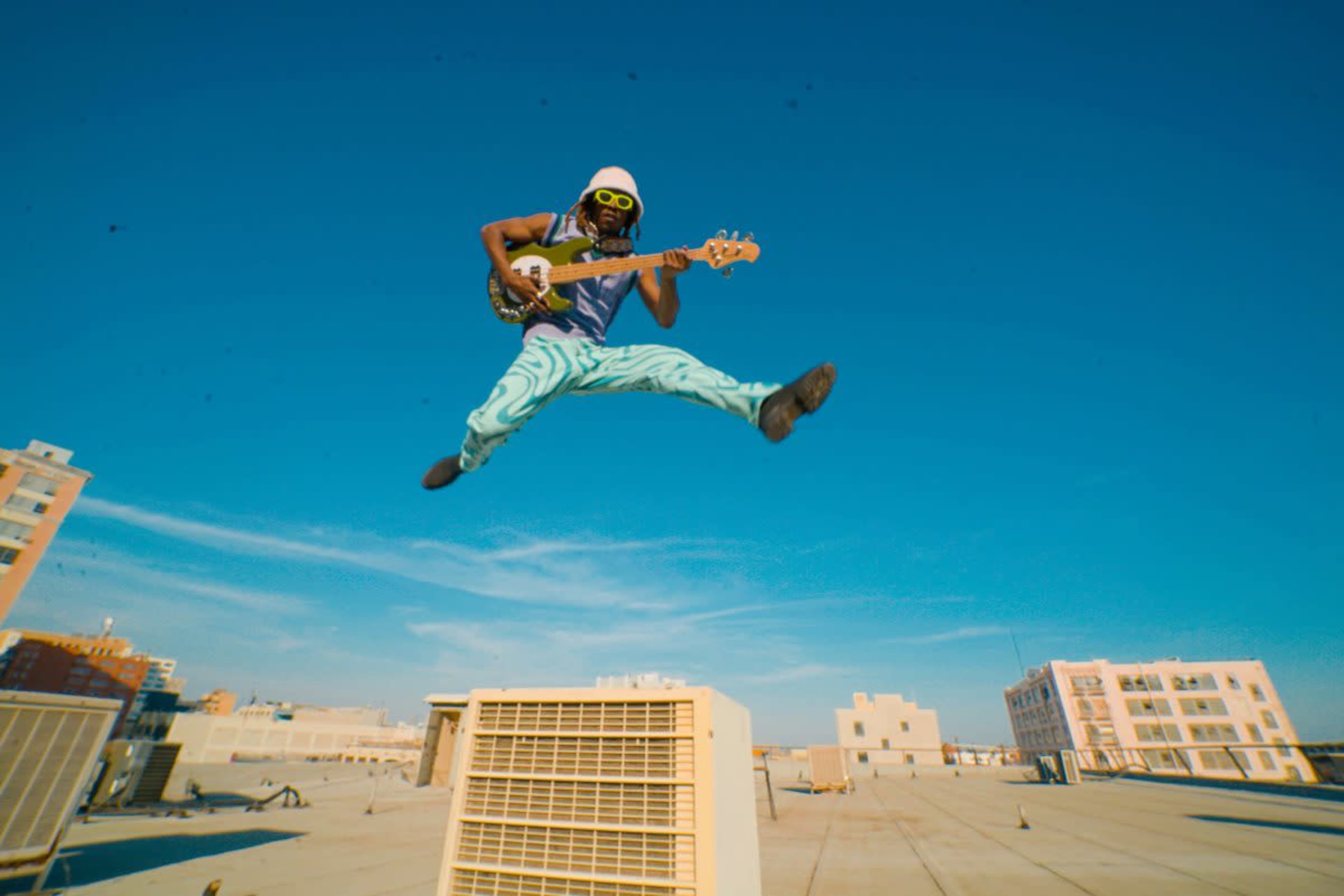 A musician in a colorful outfit jumps in the air while playing a guitar on a rooftop.