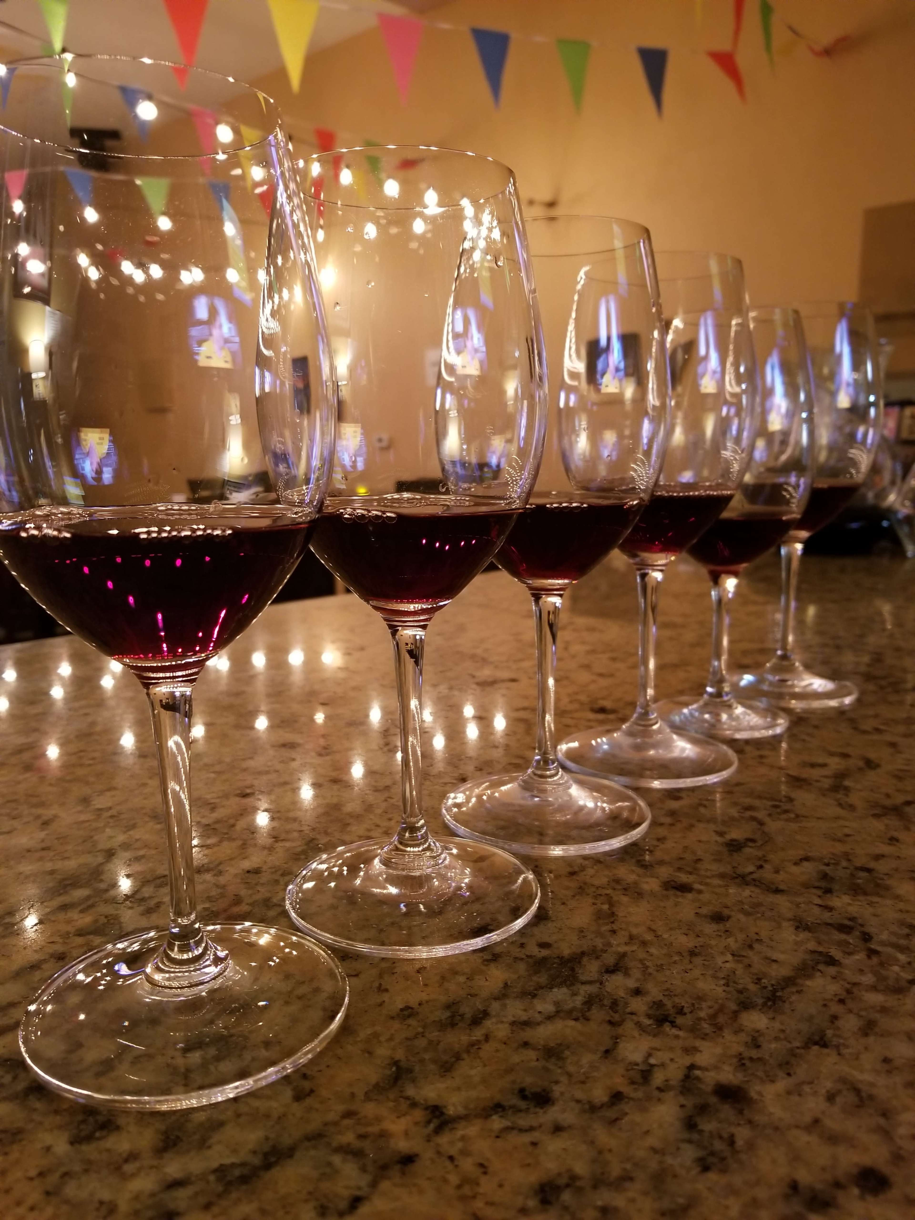 A row of wine glasses filled with red wine sits on a granite countertop under colorful party decorations.