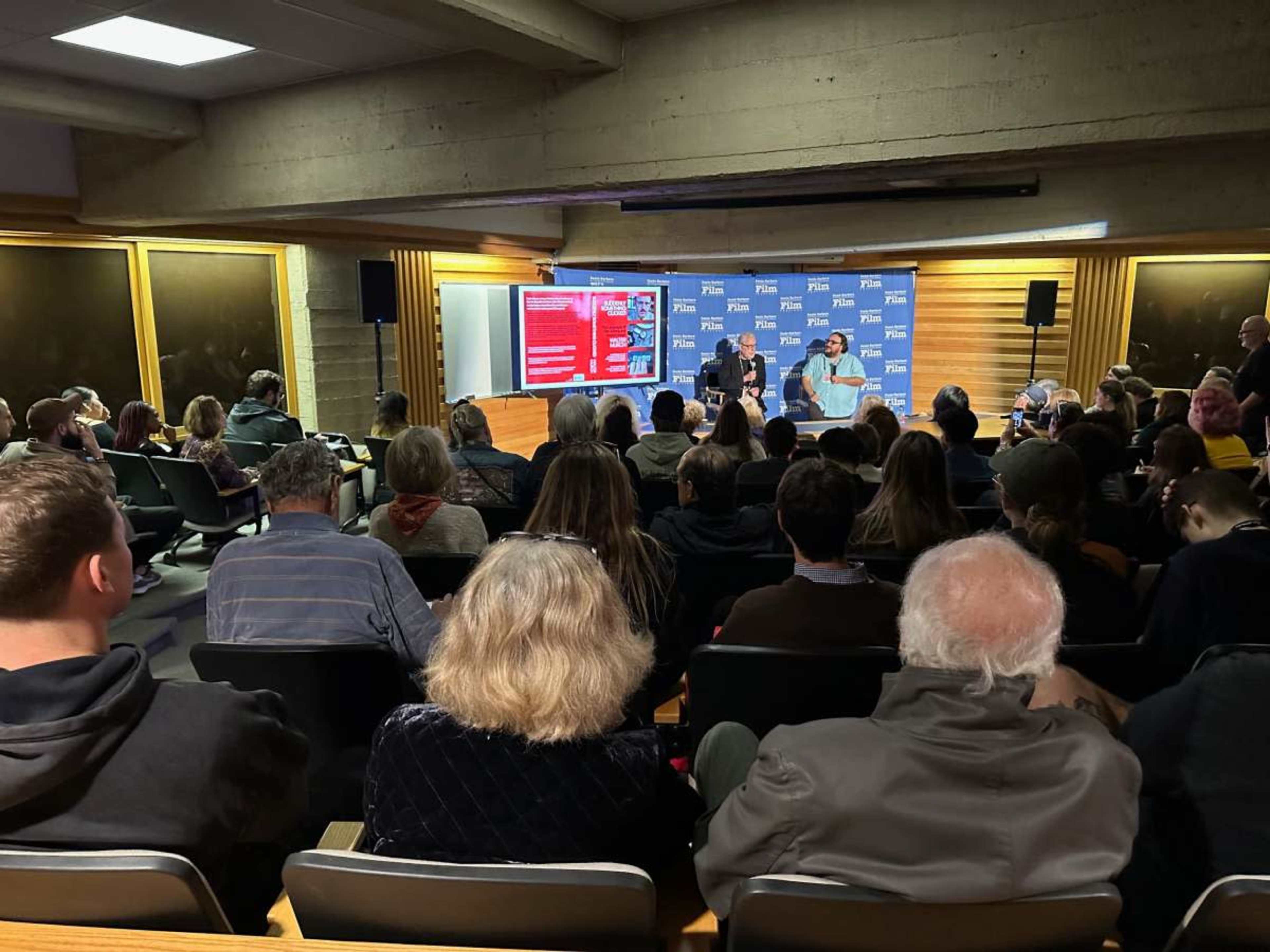 A large audience is seated in a room with a presentation area, where two speakers engage in a discussion on stage.
