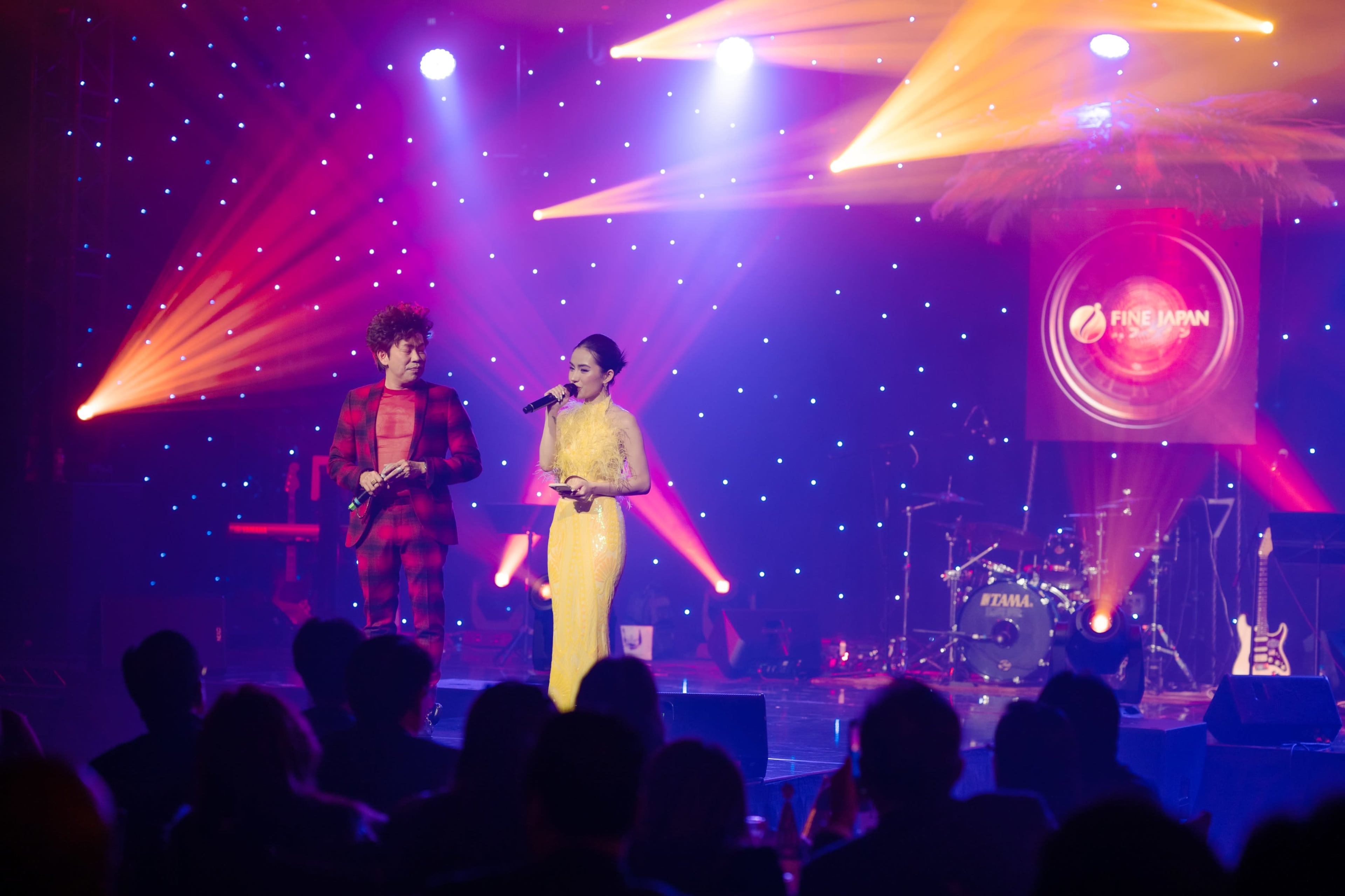 A man in a checkered suit and a woman in a yellow gown are performing on stage with colorful lights and musical instruments in the background.