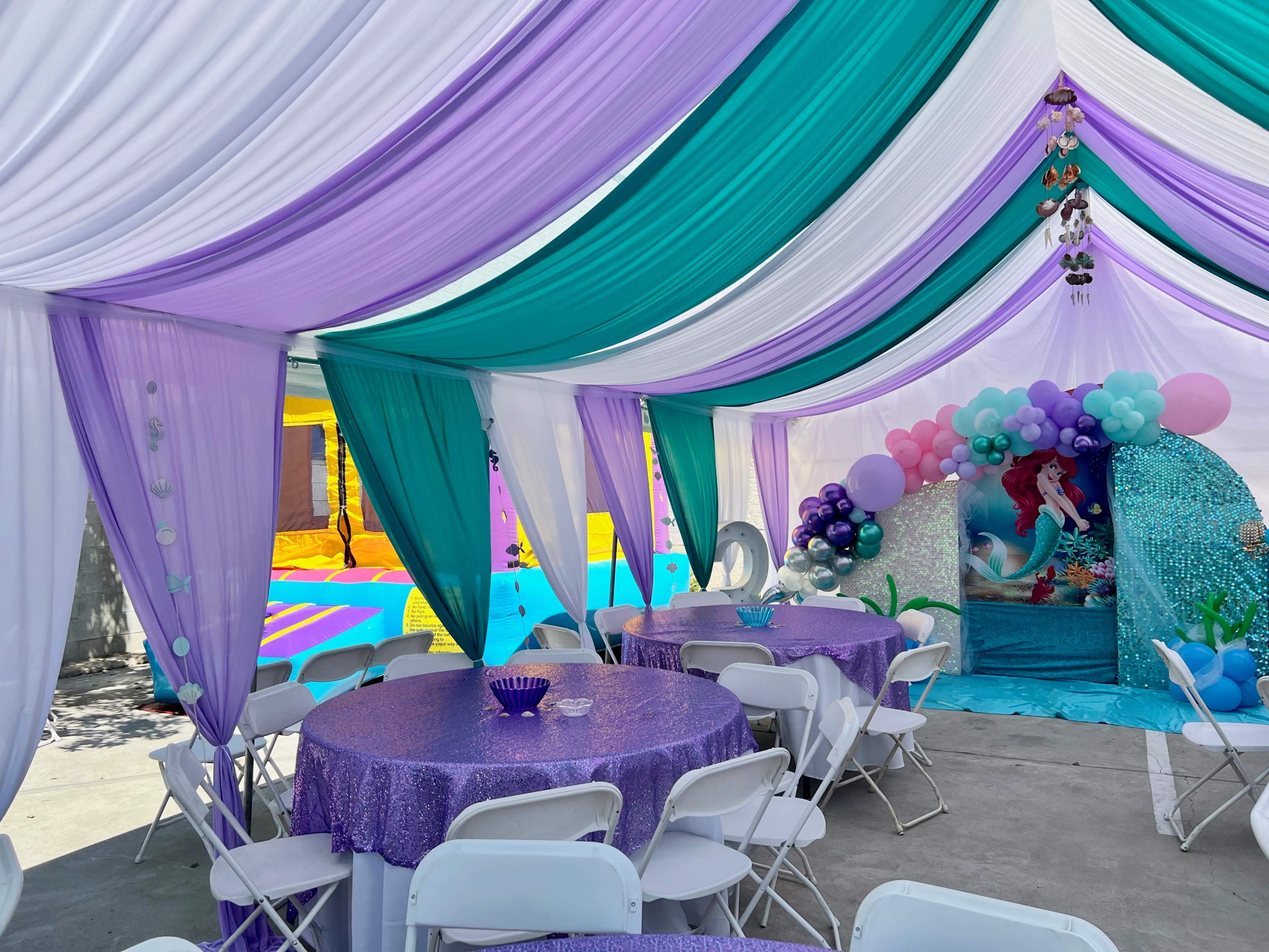 A tent decorated in purple and teal drapes houses round tables with sparkly tablecloths and features a themed backdrop with balloons.