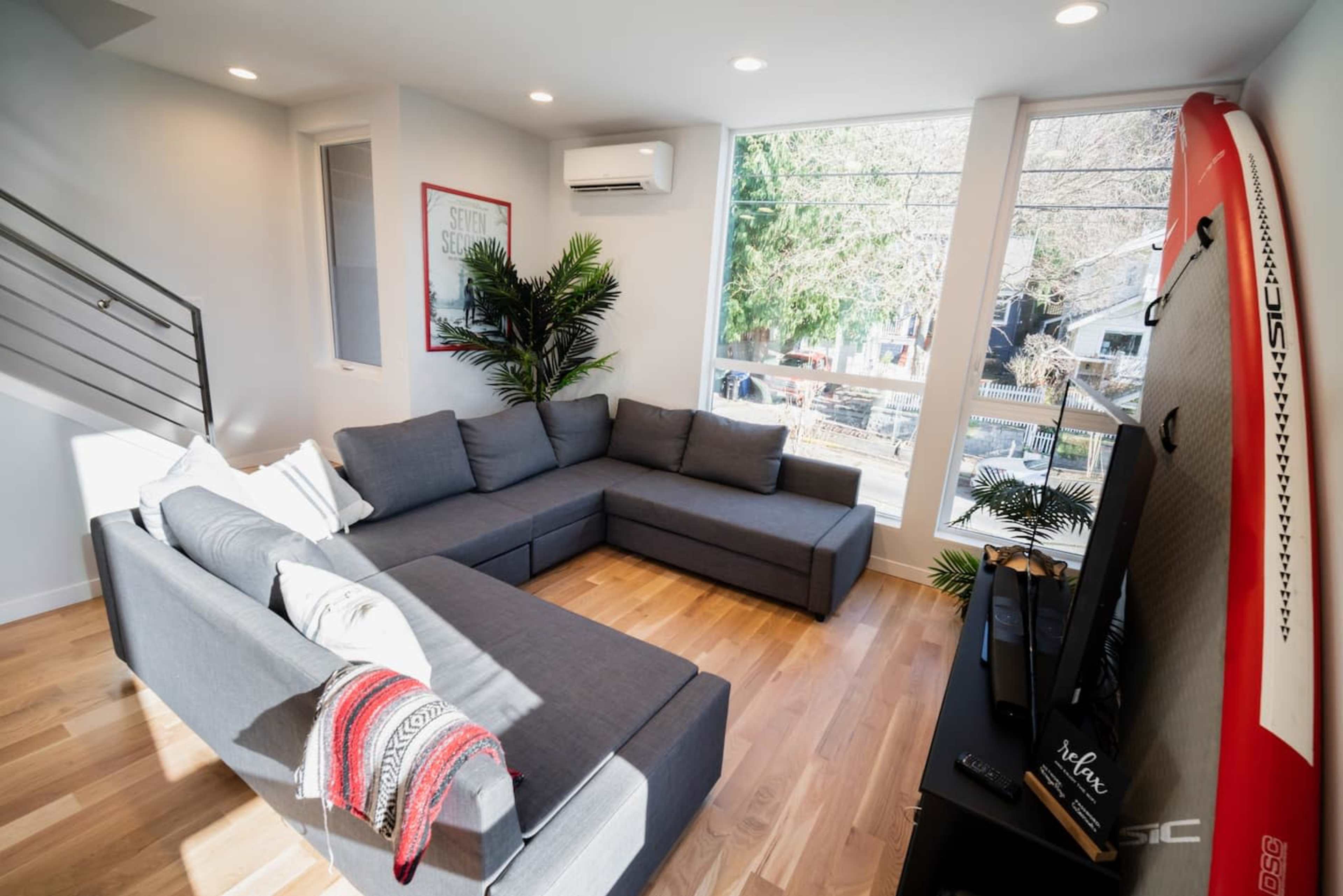 A modern living room featuring a gray sectional sofa, large windows with natural light, and a surfboard on the wall.