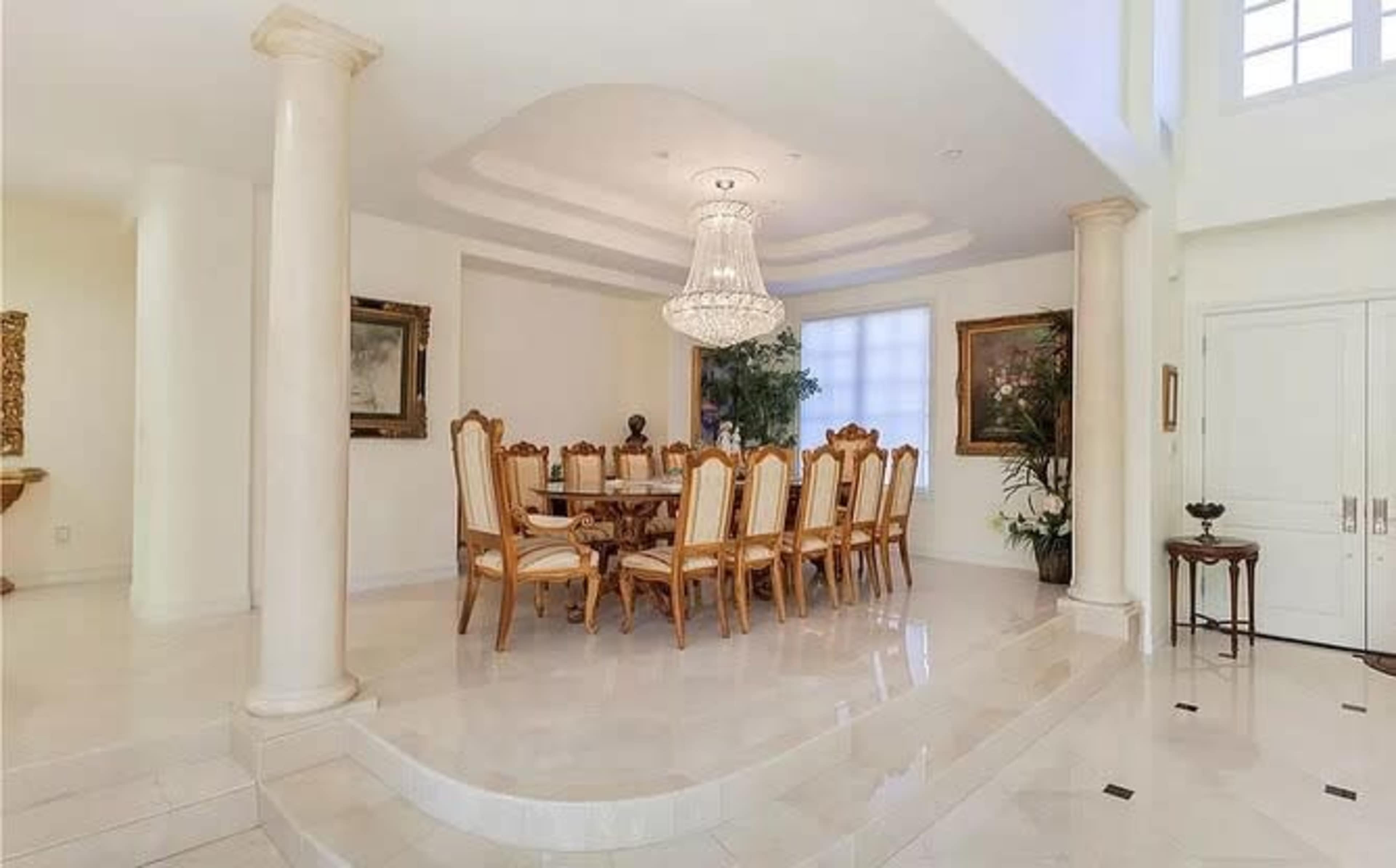 The image shows an elegant dining room with a large table surrounded by wooden chairs, a chandelier overhead, and decorative plants in the corners.