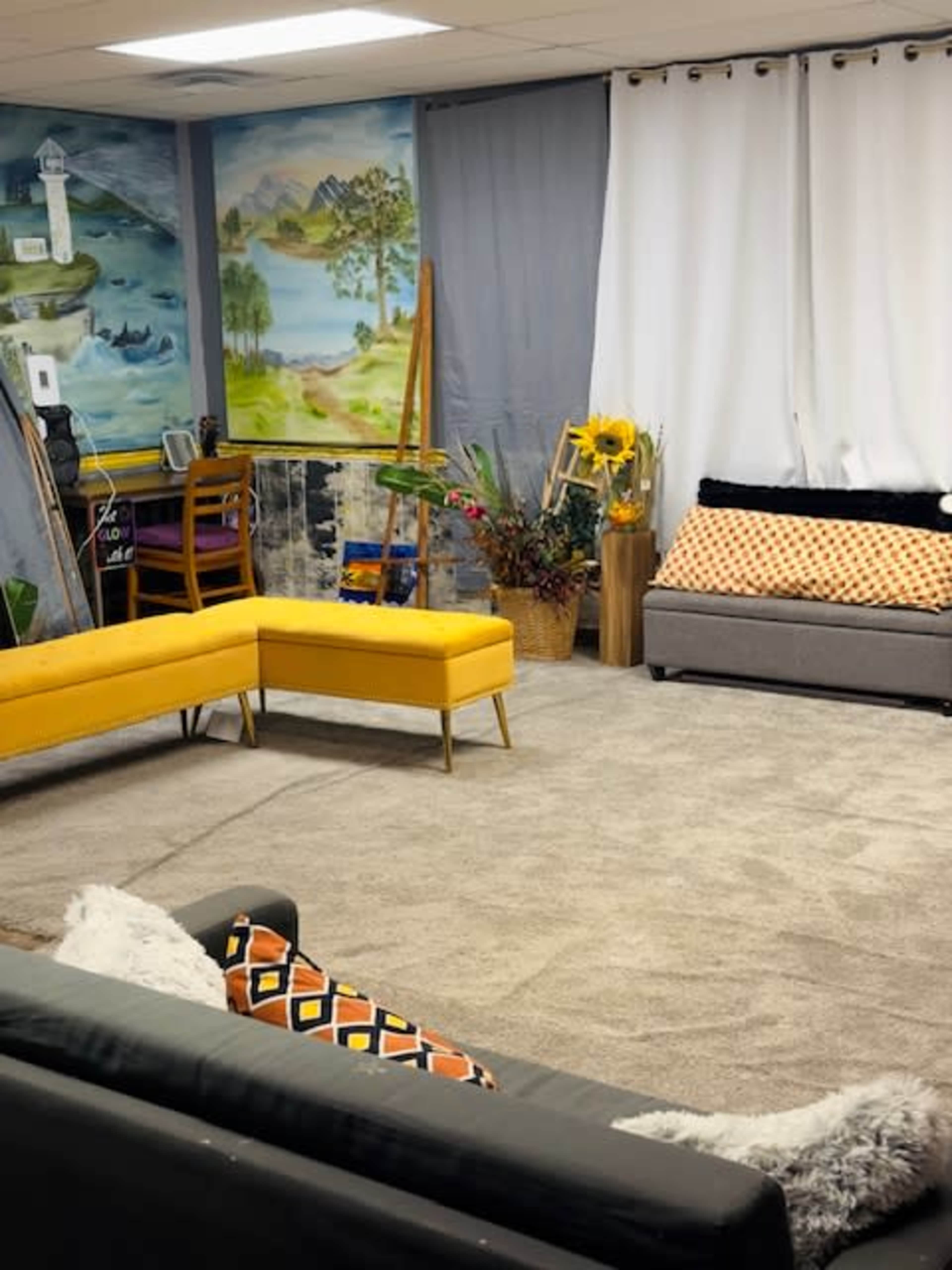 The image shows a cozy room with two sofas, a bright yellow sectional, a painting on the wall, and decorative plants.