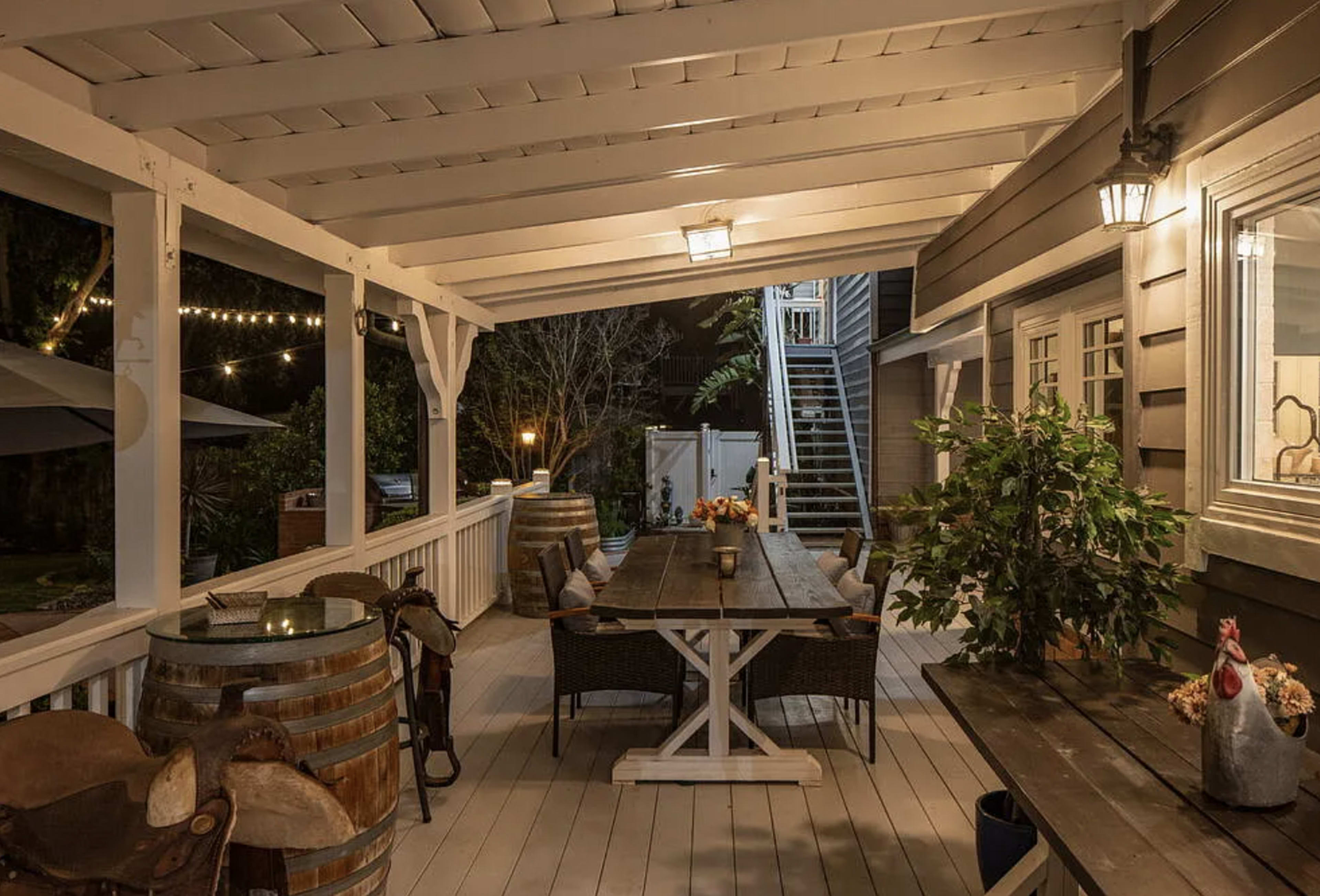 The image shows a well-lit outdoor patio with a dining table surrounded by chairs, a wooden railing, and decorative elements like plants and barrels.