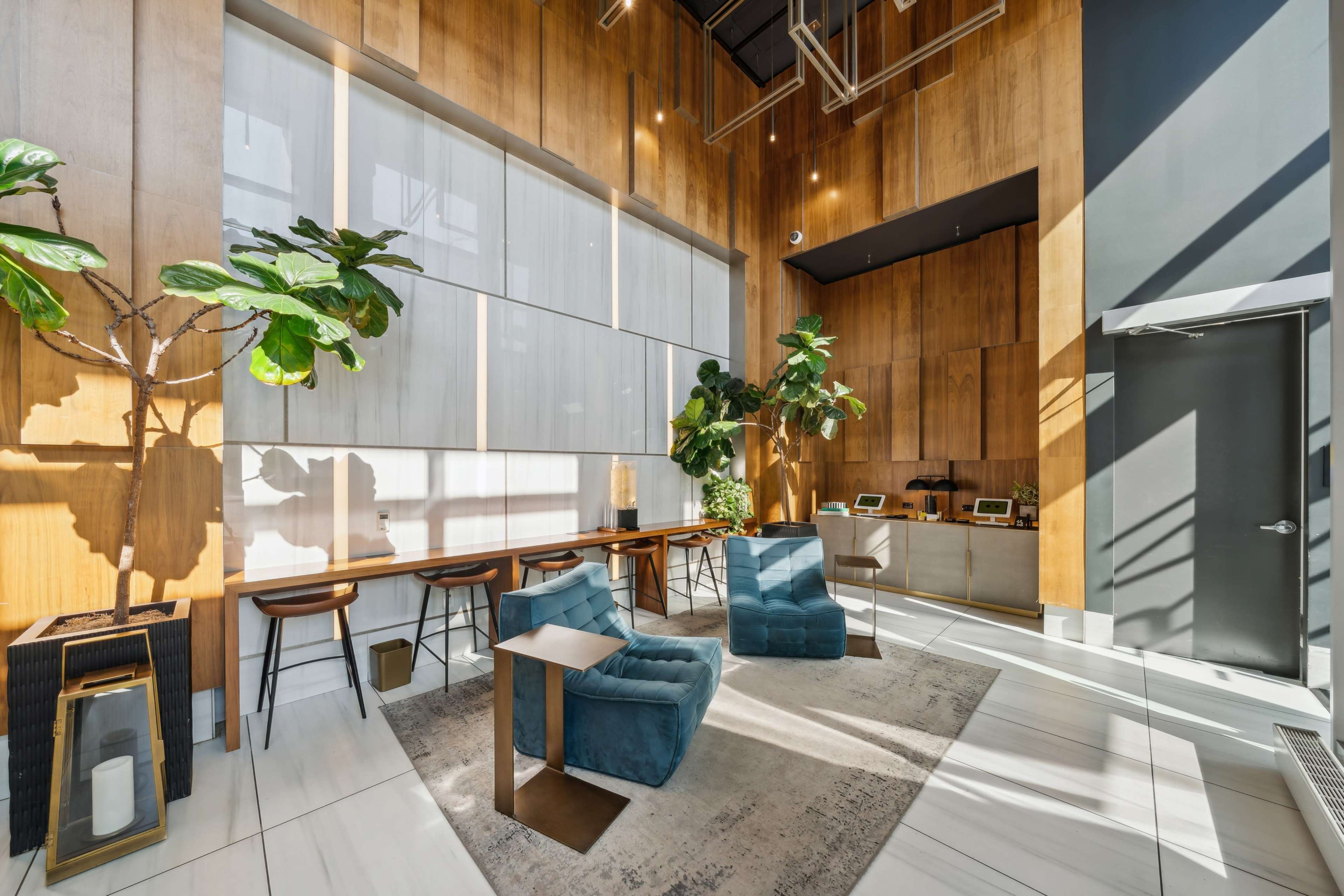 The image shows a modern lobby with wooden walls, plush blue seating, a light rug, and greenery, featuring a reception area with bar stools and a service counter.